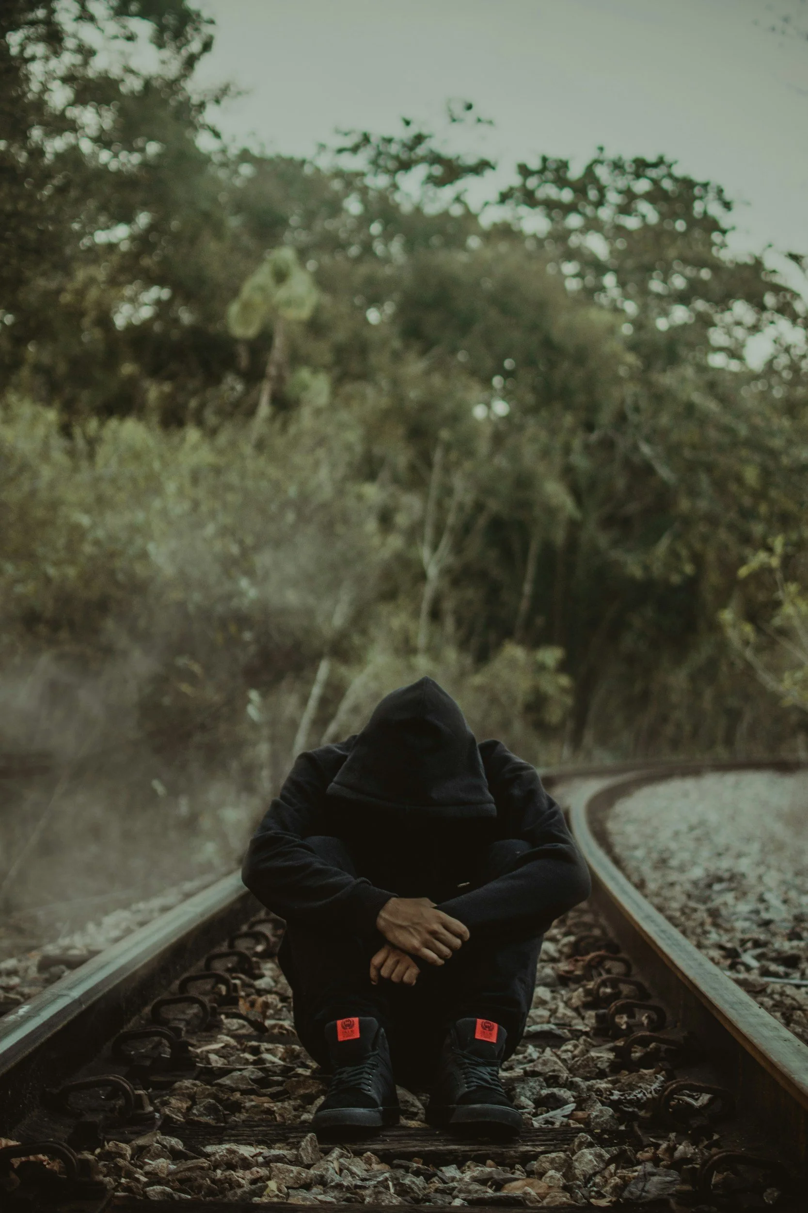 Person dressed in black sitting on train tracks with hooded hoodie, surrounded by trees and greenery.
