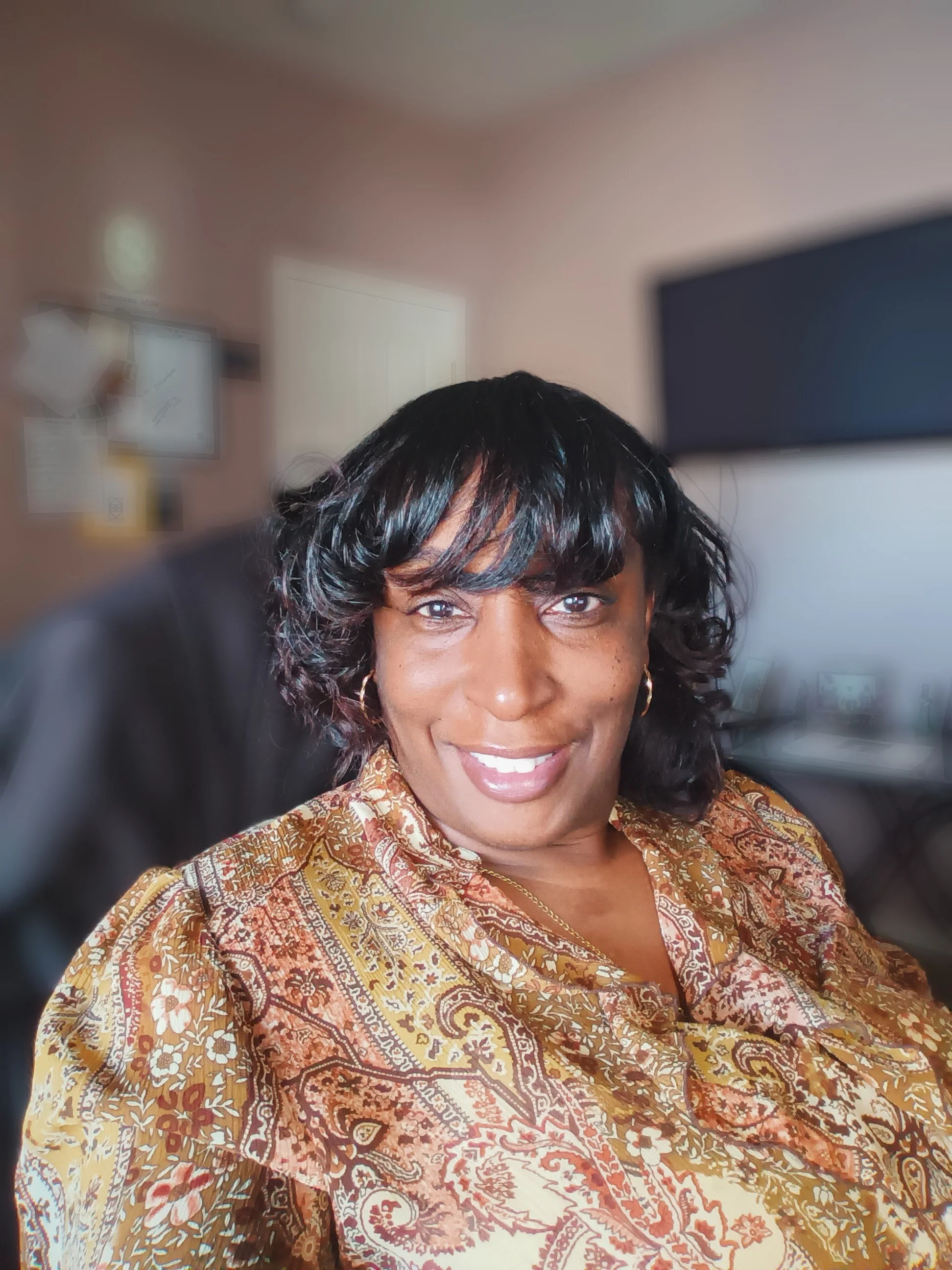 A smiling woman with dark curly hair and earrings, wearing a patterned blouse, seated in an office with a television and blurred background.