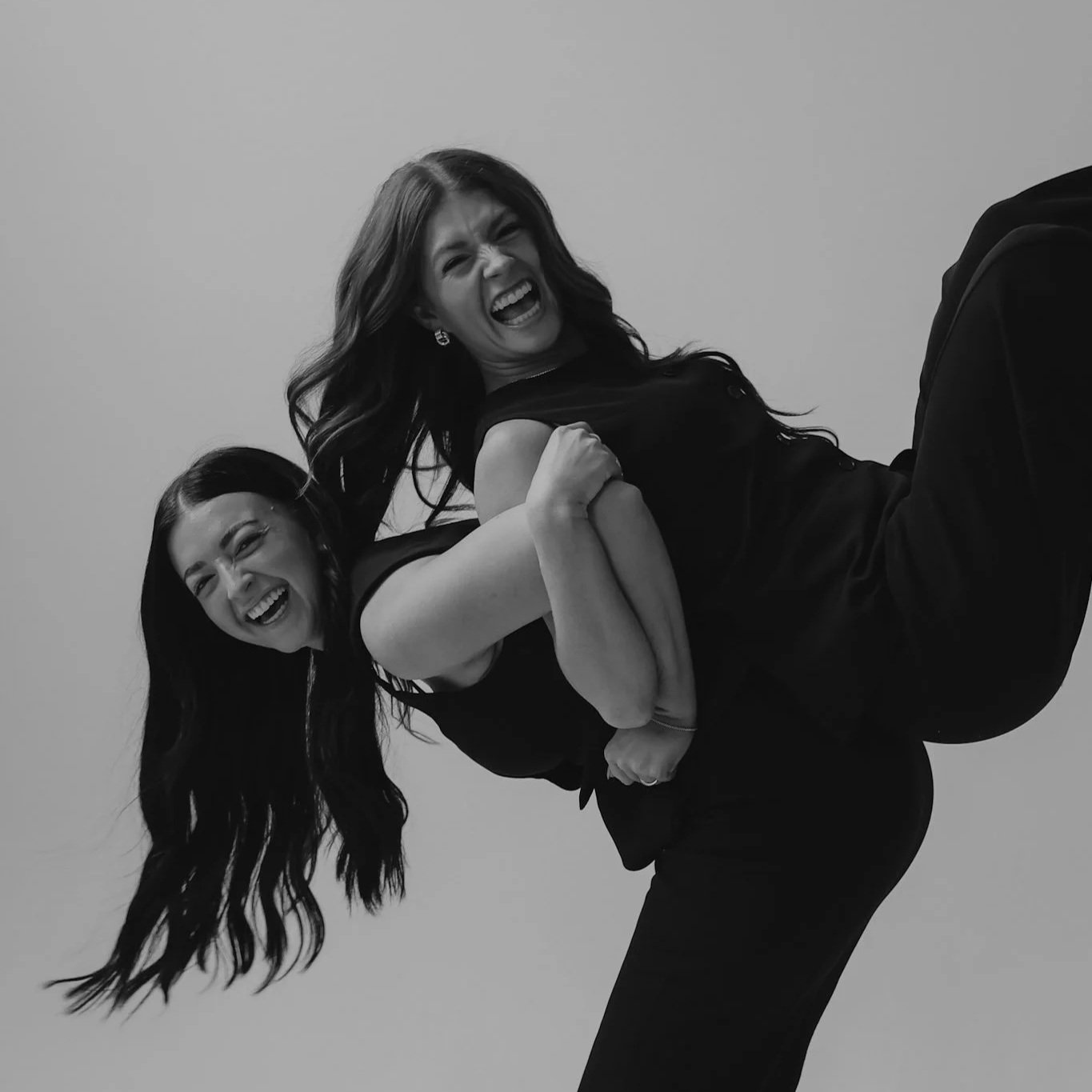 Two women with long dark hair laughing and playfully posing together, one leaning over the other's back, in black and white.