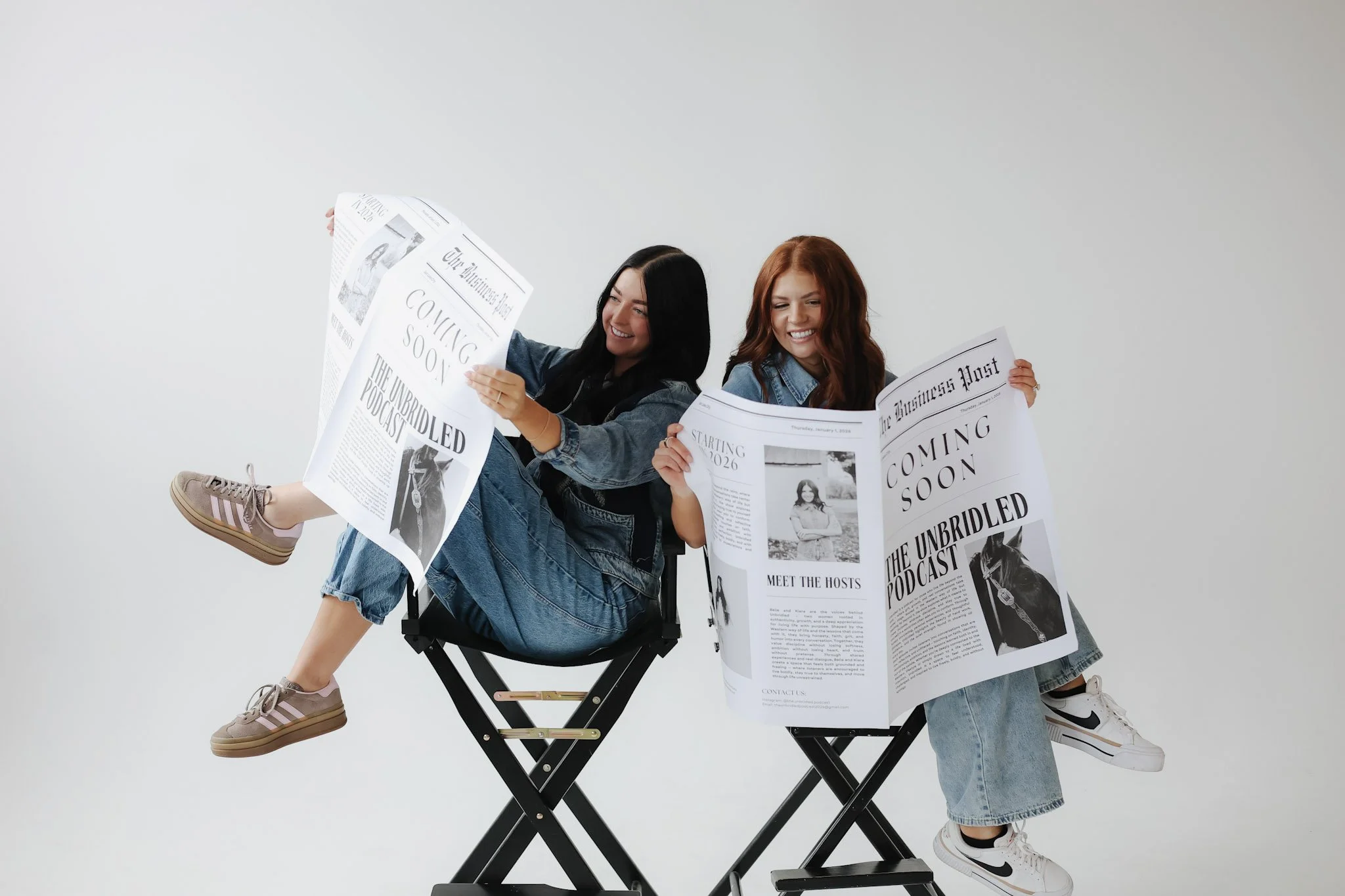 Two women sitting on director's chairs, holding newspapers and smiling.
