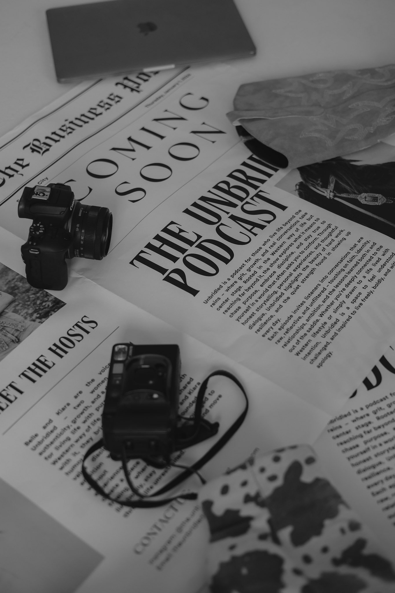 Black and white photo of a desk with newspapers, a camera, a camera lens, and a laptop.