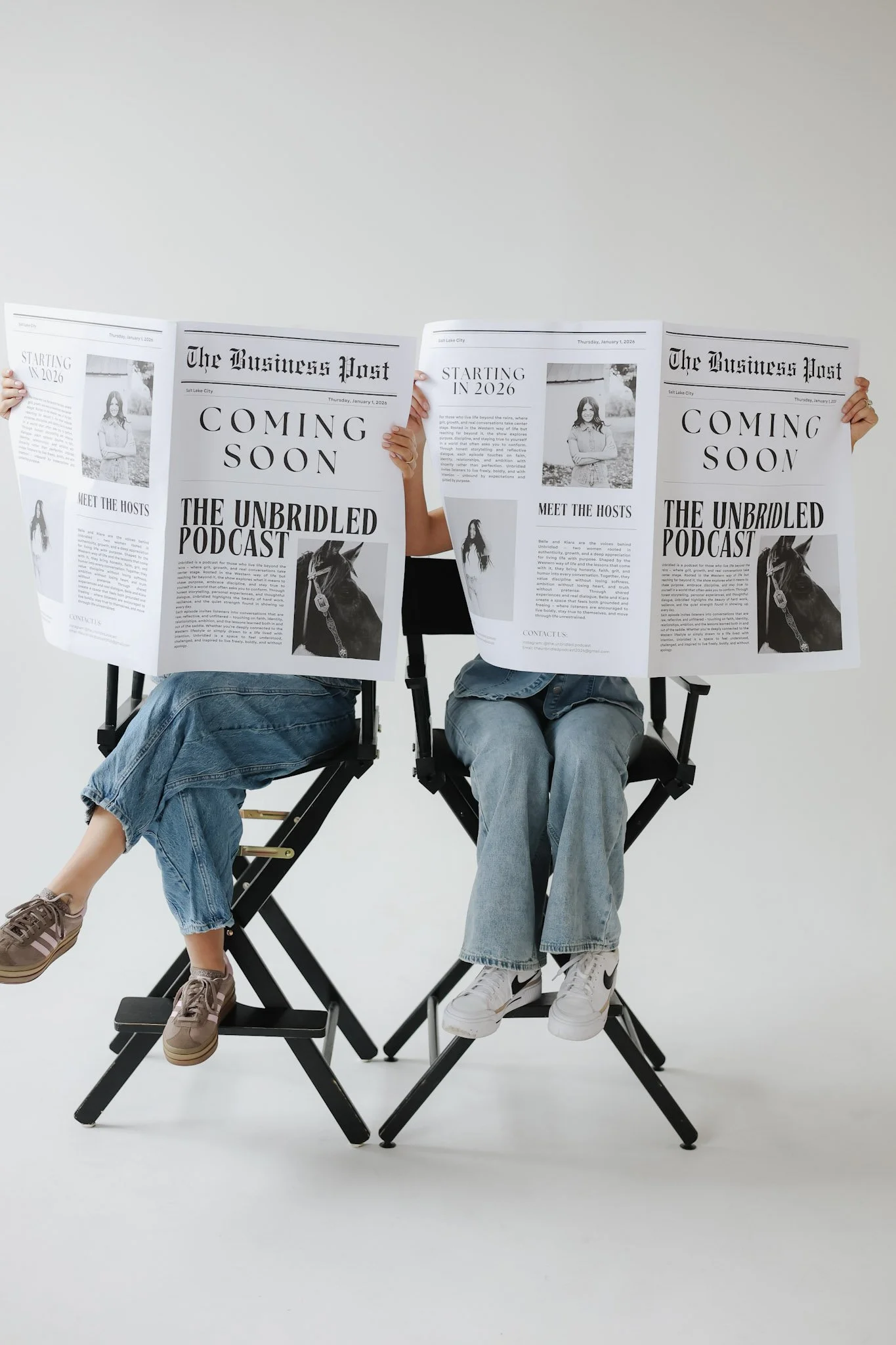 Two people sitting on black director's chairs with large newspapers covering their faces, in a photo studio with a plain white background.