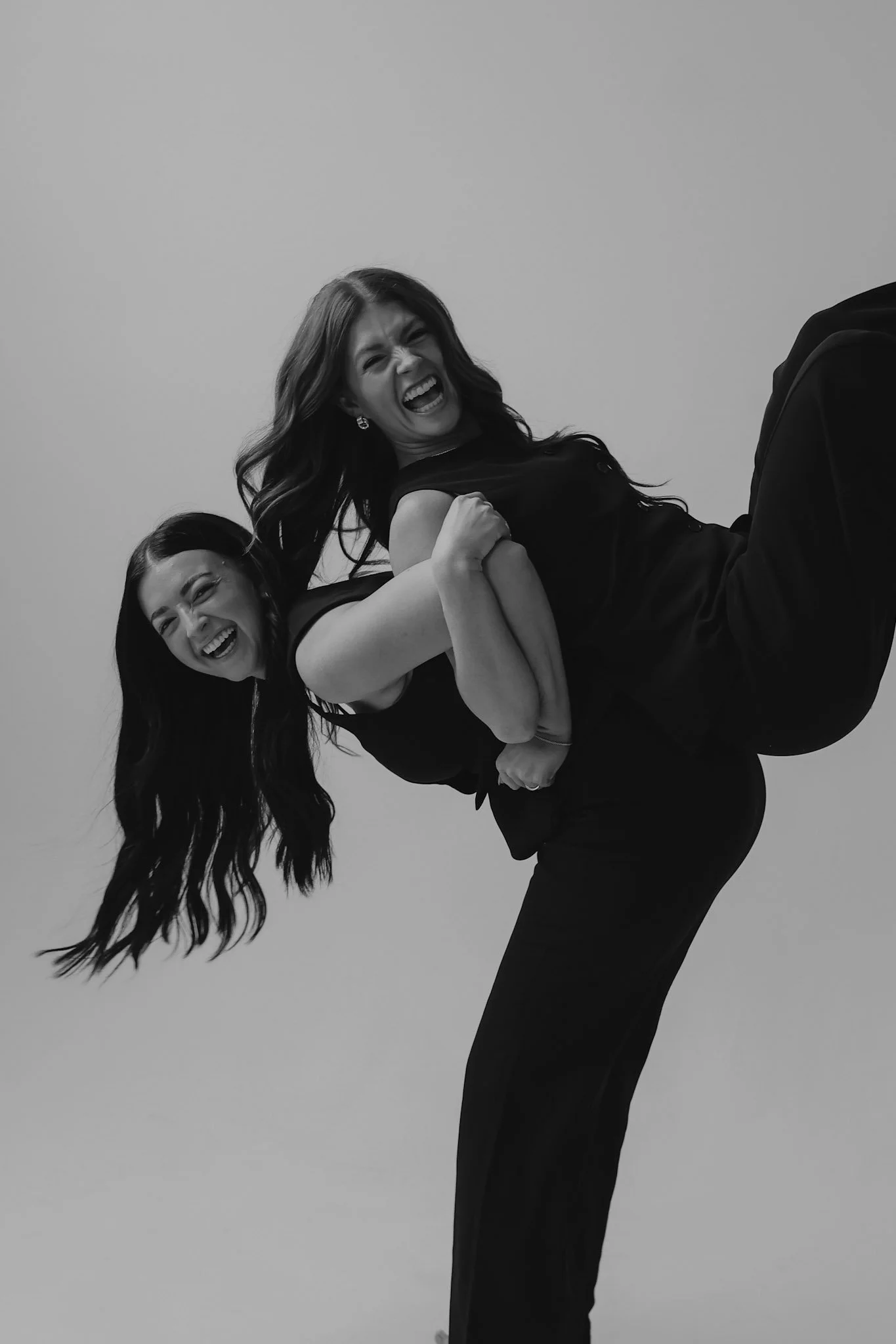 Two women in black dresses are smiling and laughing, one is holding the other upside down in a playful pose against a plain background.