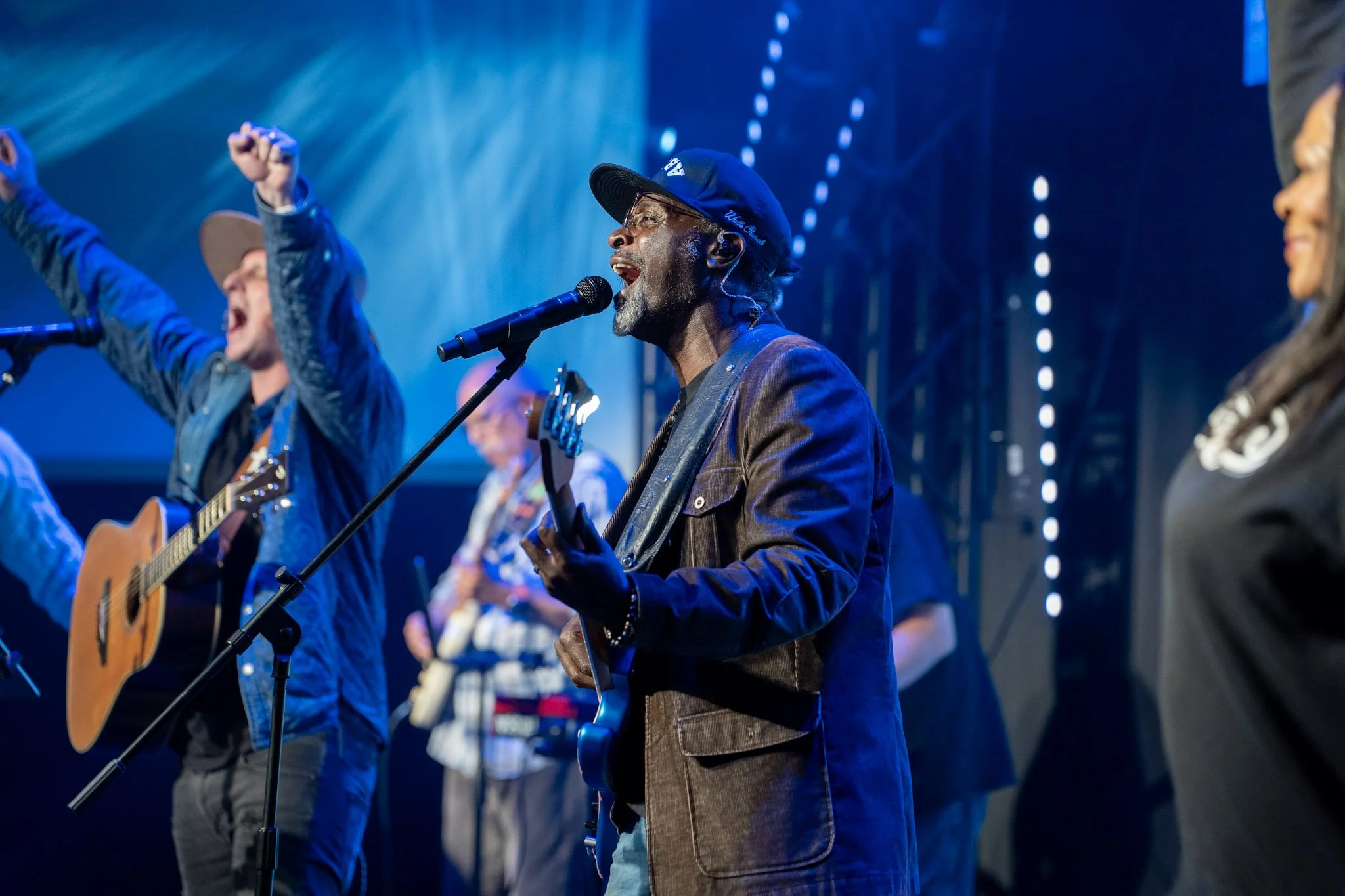 Group of musicians performing live on stage, singing and playing guitars, with stage lighting in the background.