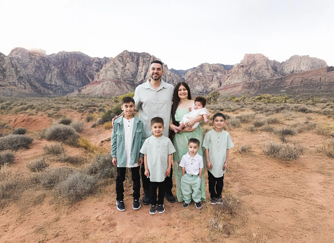 A family of eight standing on a dirt trail in a desert landscape with mountainous terrain in the background. The adults are holding a baby girl, and all are smiling at the camera.