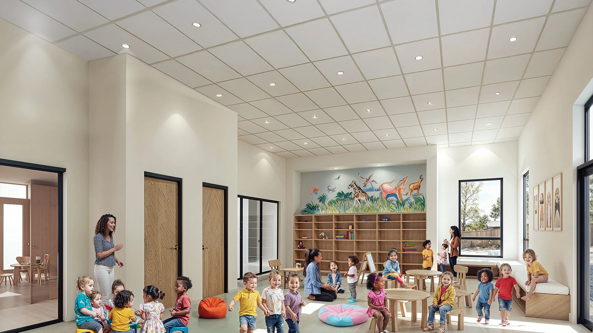 Children and teachers in a bright, spacious indoor playroom with large windows, wooden tables, a bookshelf, and colorful wall art of animals and nature.