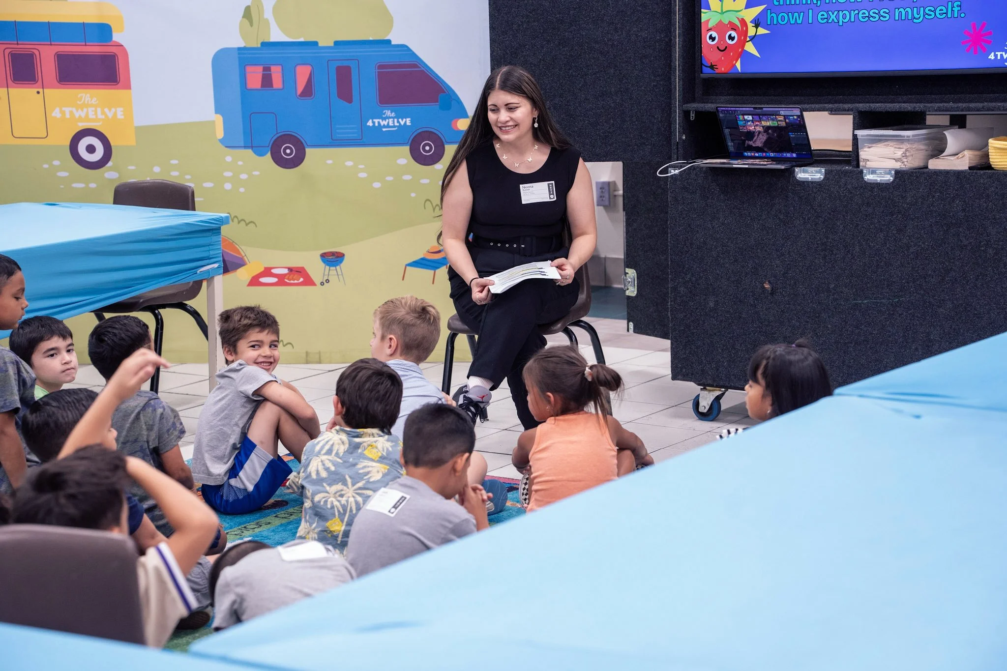 A woman sitting on a chair in front of a group of children, holding a book and smiling. The children are sitting on the floor, some looking at her, in a colorful classroom with a mural of trucks and outdoor scene on the wall.