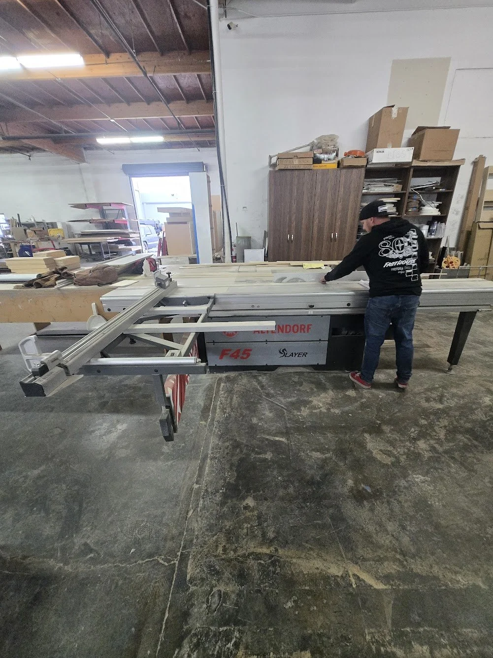 A person working with a sliding table saw in a woodworking shop, surrounded by shelves and supplies.