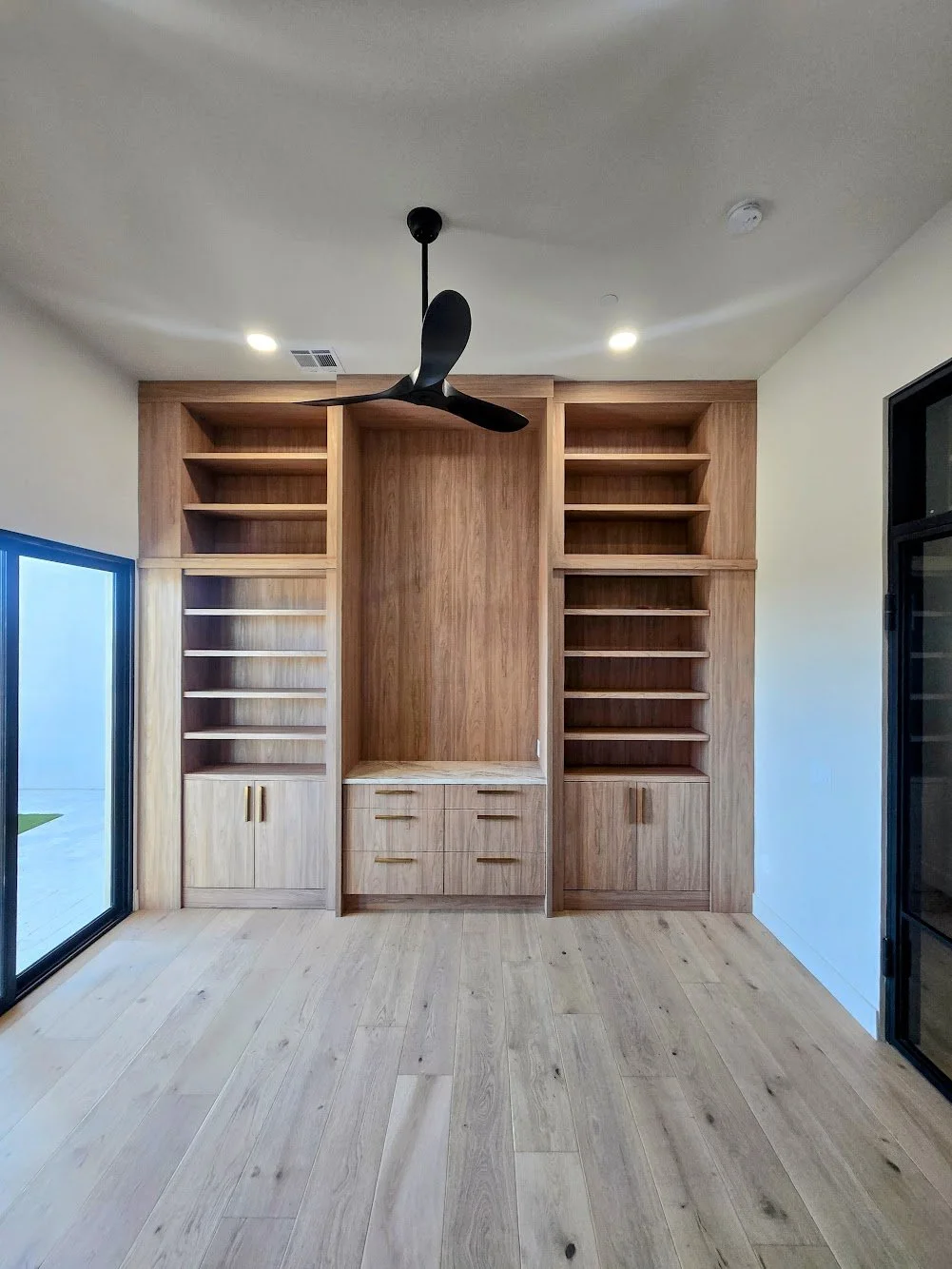 Empty built-in wooden shelving unit with drawers in a modern room, ceiling fan, and sliding glass door.