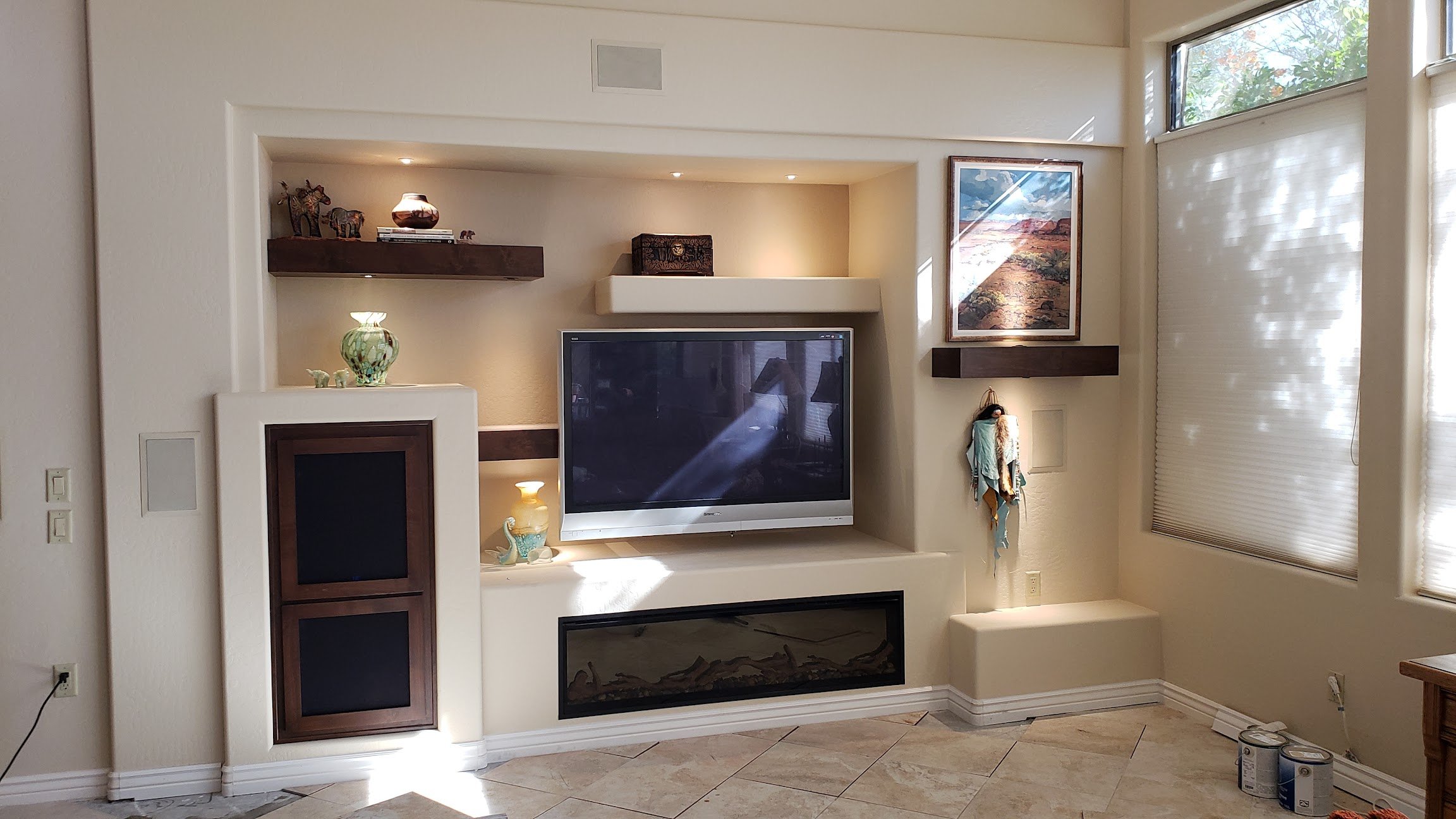 Living room with an entertainment center, featuring a large flat-screen TV, decorative shelves with artwork and ornaments, a framed landscape painting, and a window with blinds letting in natural light.