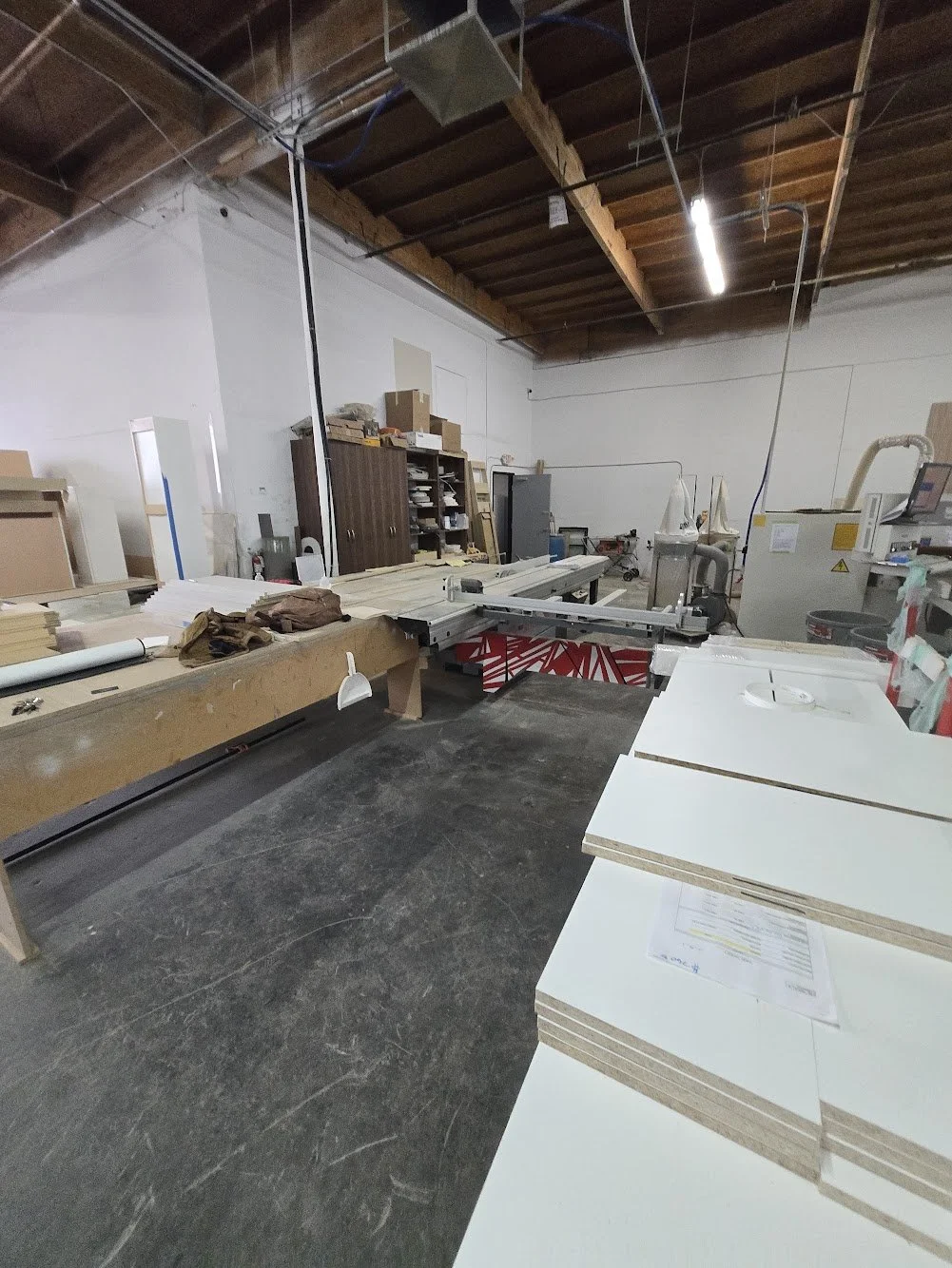 Workshop with woodworking equipment, including a large table saw, stacks of MDF boards, and various tools and supplies, with wooden ceiling beams and exposed wiring.