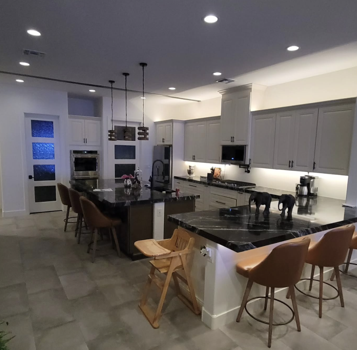 Modern kitchen with white cabinets, black marble countertops, and a kitchen island with four chairs. There are two black dogs on the counter, and various appliances and decor items are visible. The space is well-lit with recessed ceiling lights.