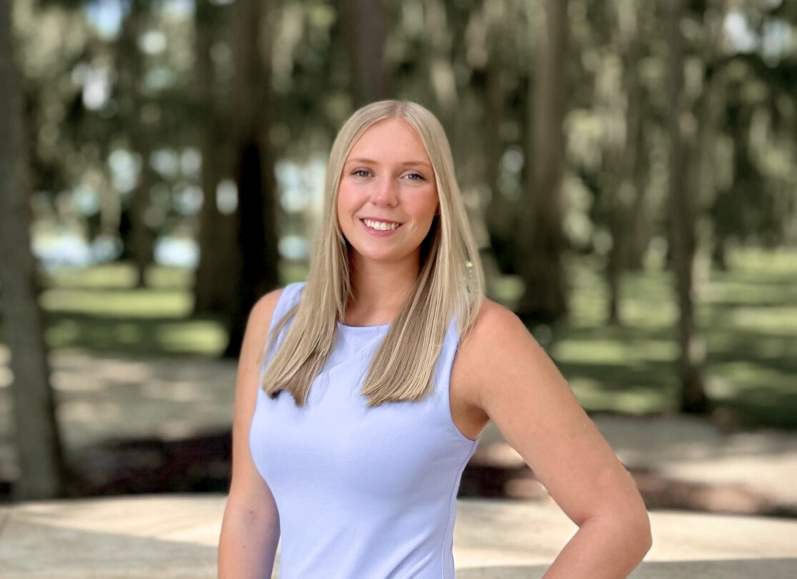 A smiling blonde woman in a sleeveless light blue top standing outdoors in a wooded park.