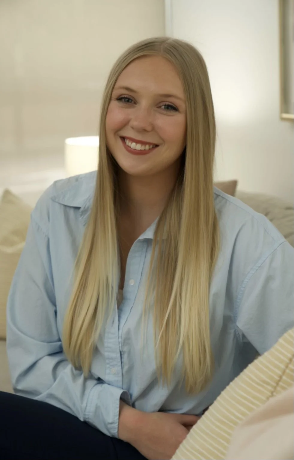 A young woman with long blonde hair, smiling, sitting indoors in a light blue shirt.
