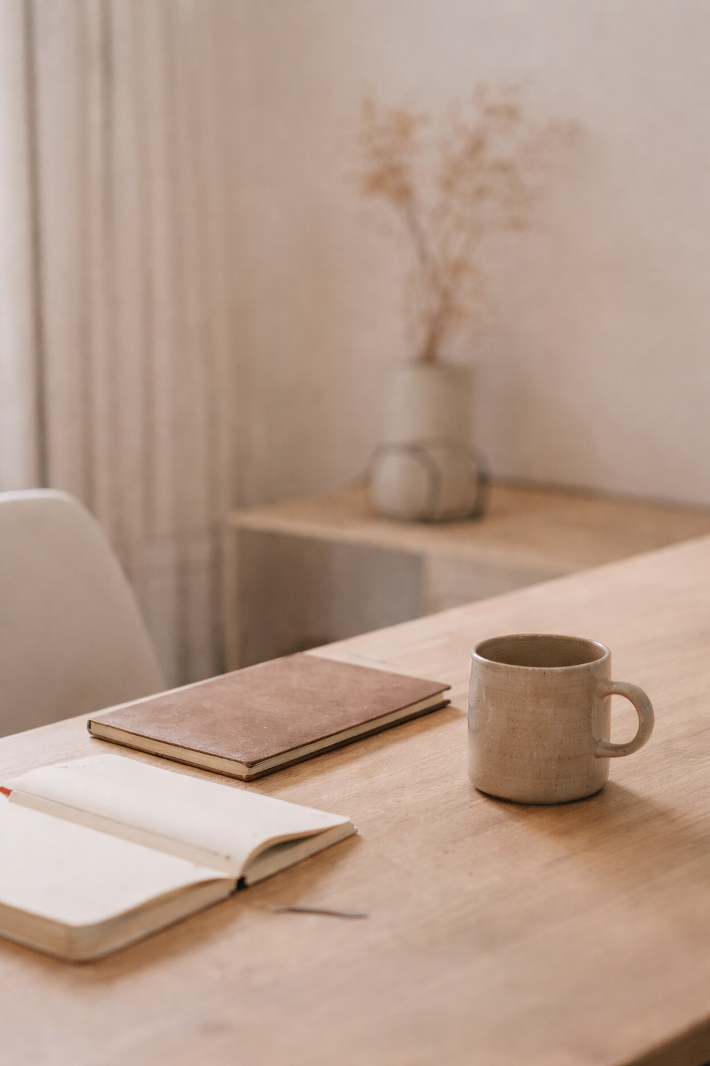 A ceramic mug, two notebooks, and a pen arranged on a wooden table in a cozy, well-lit room.