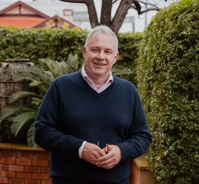 A smiling man with gray hair in a navy sweater standing outdoors in a garden with green bushes and trees.