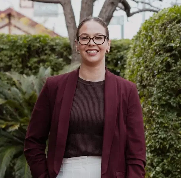A woman smiling outdoors, wearing glasses, a maroon blazer, a dark brown top, and light-colored pants, with greenery and trees in the background.
