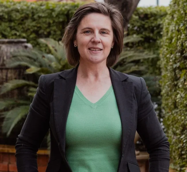A woman in a black blazer and green shirt standing outdoors with greenery in the background.
