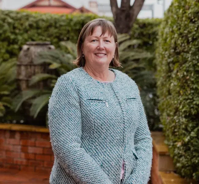 A woman with short brown hair standing outdoors in a garden, smiling, dressed in a light blue textured blazer.