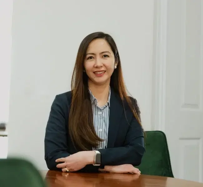 A woman with long brown hair, wearing a dark blazer and striped shirt, sitting at a wooden table in a room with white walls and a closed door.