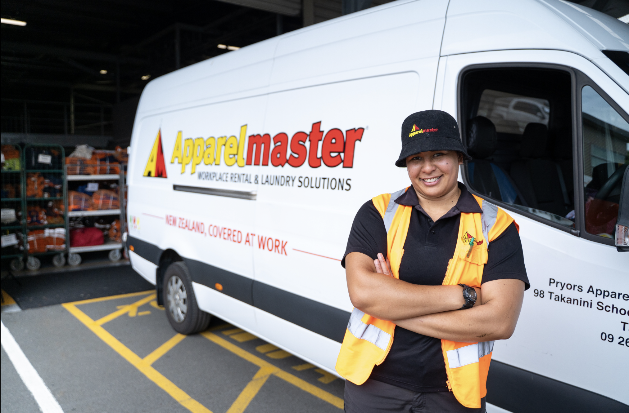 A woman in a black shirt and safety vest smiling with her arms crossed, standing in front of a white company van with logo and text, in a parking lot near shelving with orange work supplies.