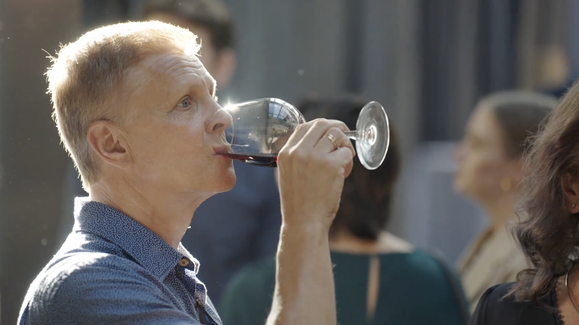 A woman with short blonde hair and fair skin is drinking red wine from a wine glass at an event, with other people in the background.