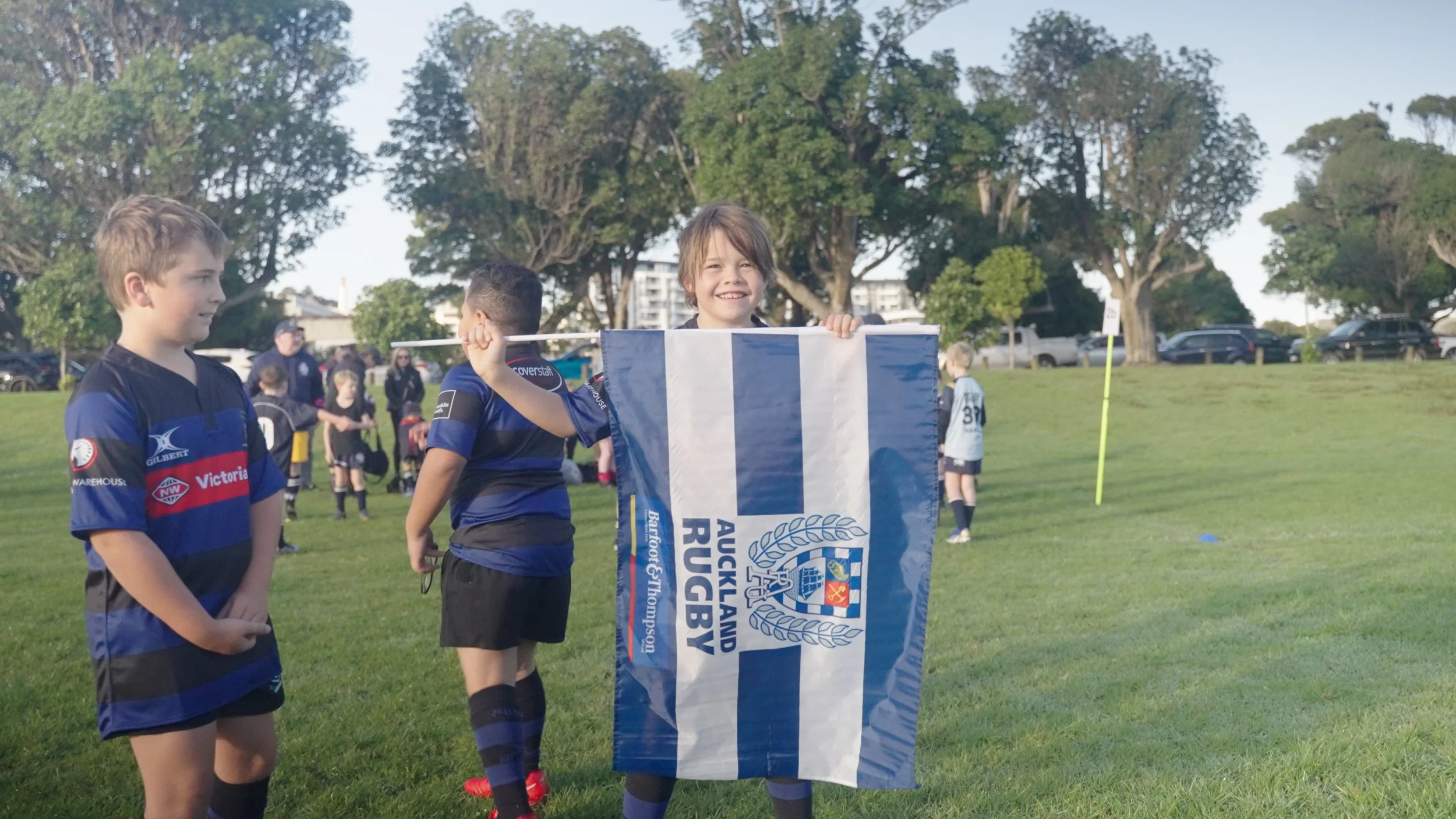 Young boys in rugby uniforms on a grassy field, one boy holding an Auckland Rugby flag and smiling at the camera.