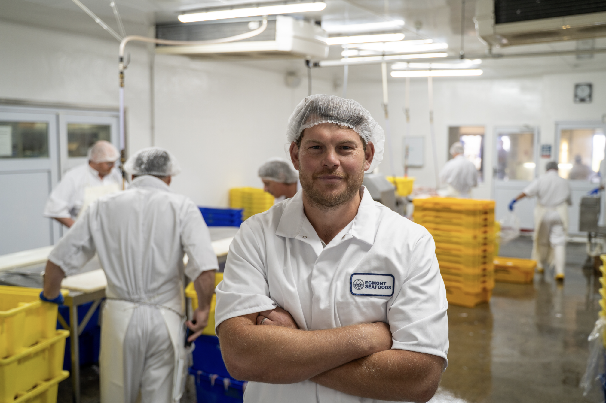 A man with a beard wearing a hairnet and white apron with a name tag that reads 'EGMONT SEAFOODS' standing with arms crossed inside a seafood processing facility with other workers in the background.