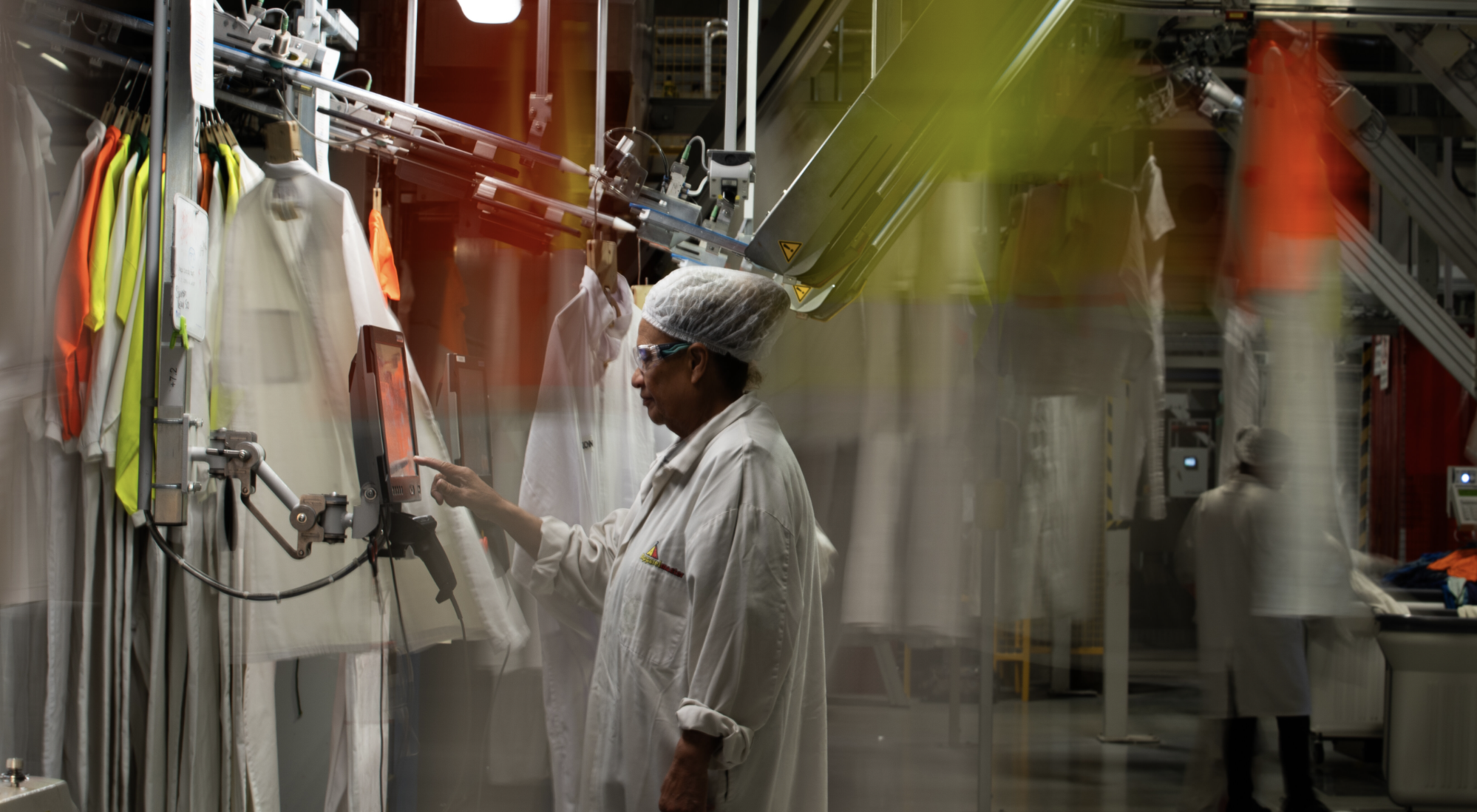 A worker in a white lab coat, hairnet, and safety glasses operates a touchscreen monitor inside a garment manufacturing or laboratory environment. There are hanging white lab coats or protective clothing on the left, and automated machinery in the ba
