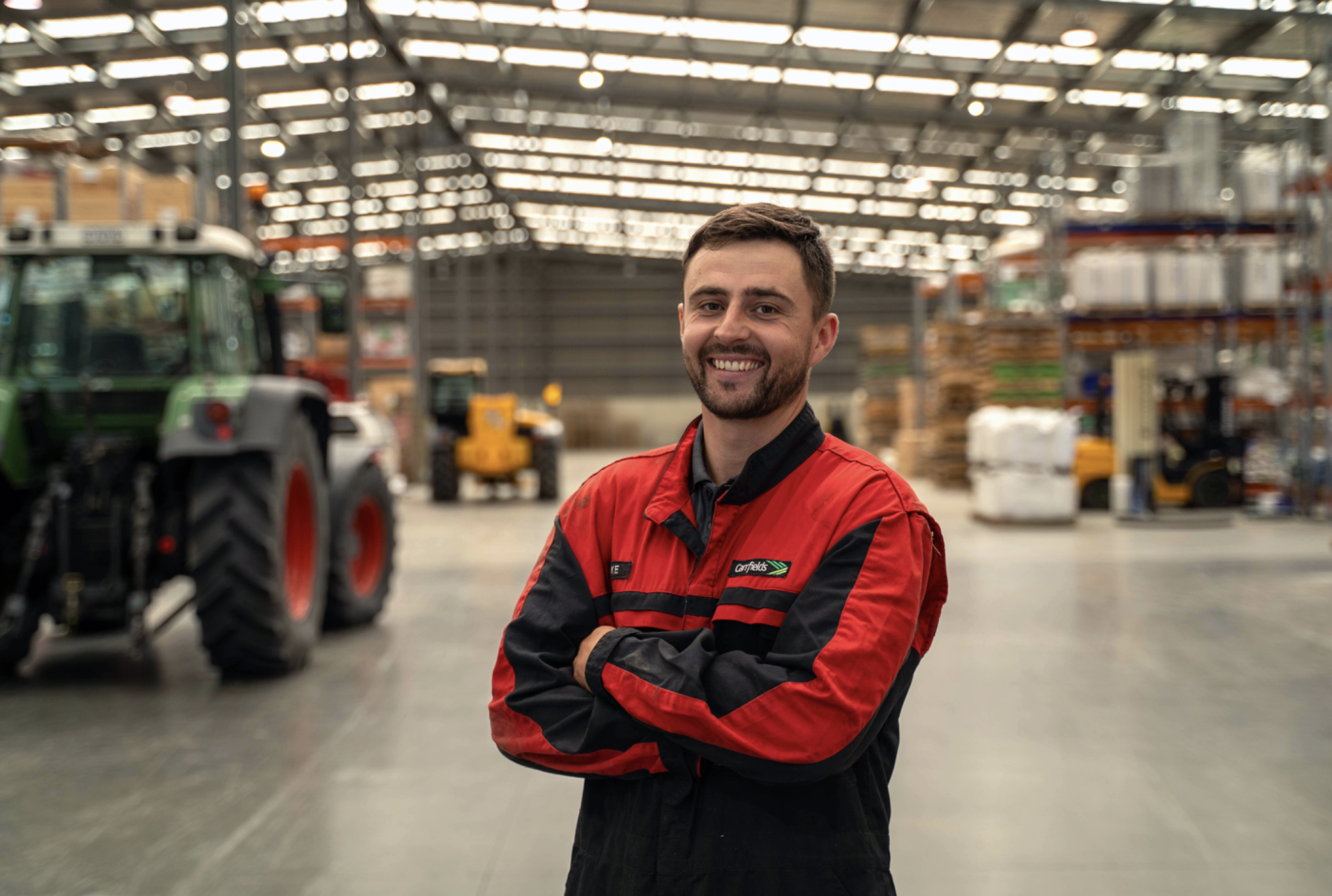 A man smiling with arms crossed, wearing a red and black uniform, standing inside a warehouse with machinery and shelves stacked with goods.