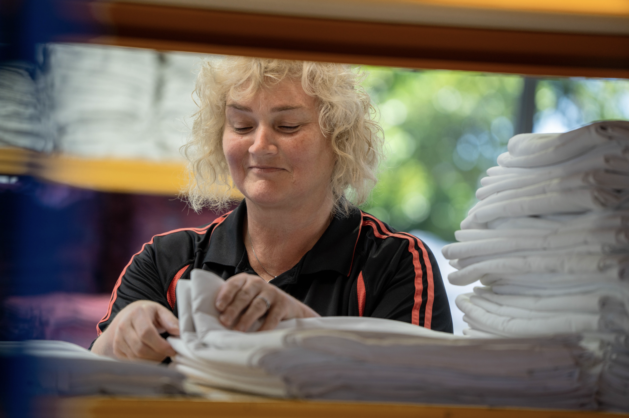 A woman with curly blonde hair looking at papers on a table, surrounded by stacks of papers and folders, with a window showing green foliage in the background.
