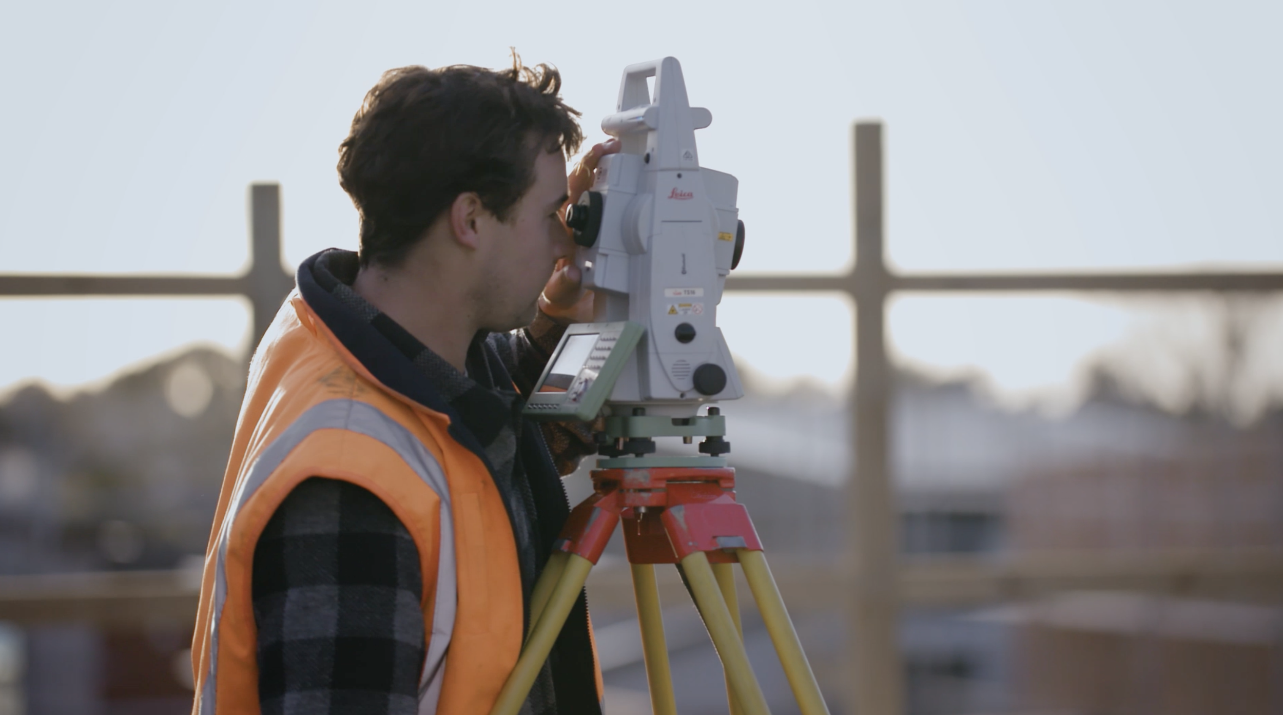 A surveyor in an orange safety vest operates a theodolite or total station device on a construction site, with a blurred background of fencing and construction materials.