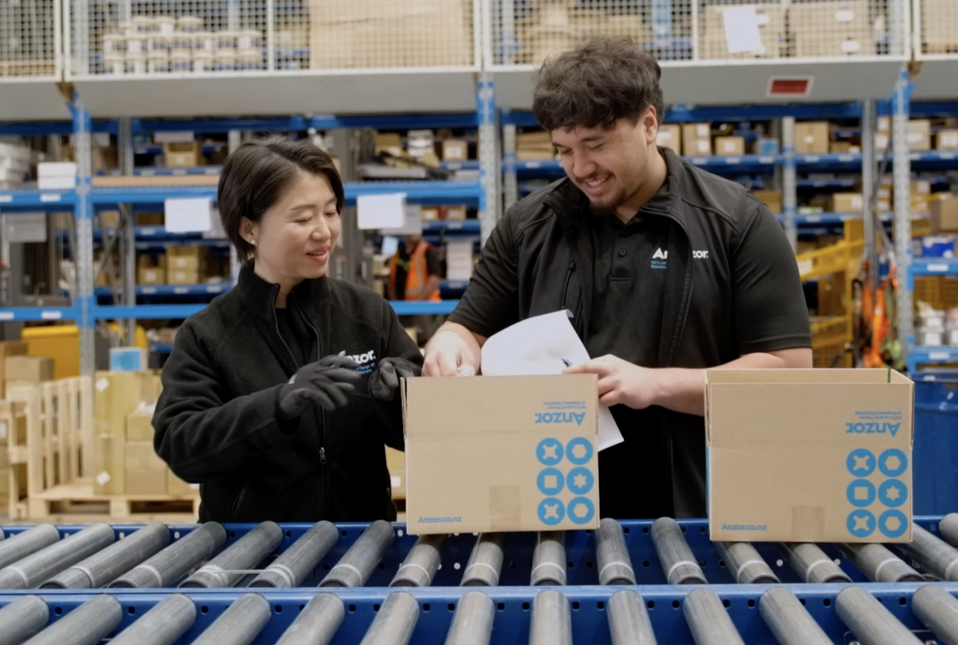Two warehouse workers, a woman and a man, are smiling and inspecting a cardboard box on a conveyor belt in a storage facility filled with shelves of boxes.