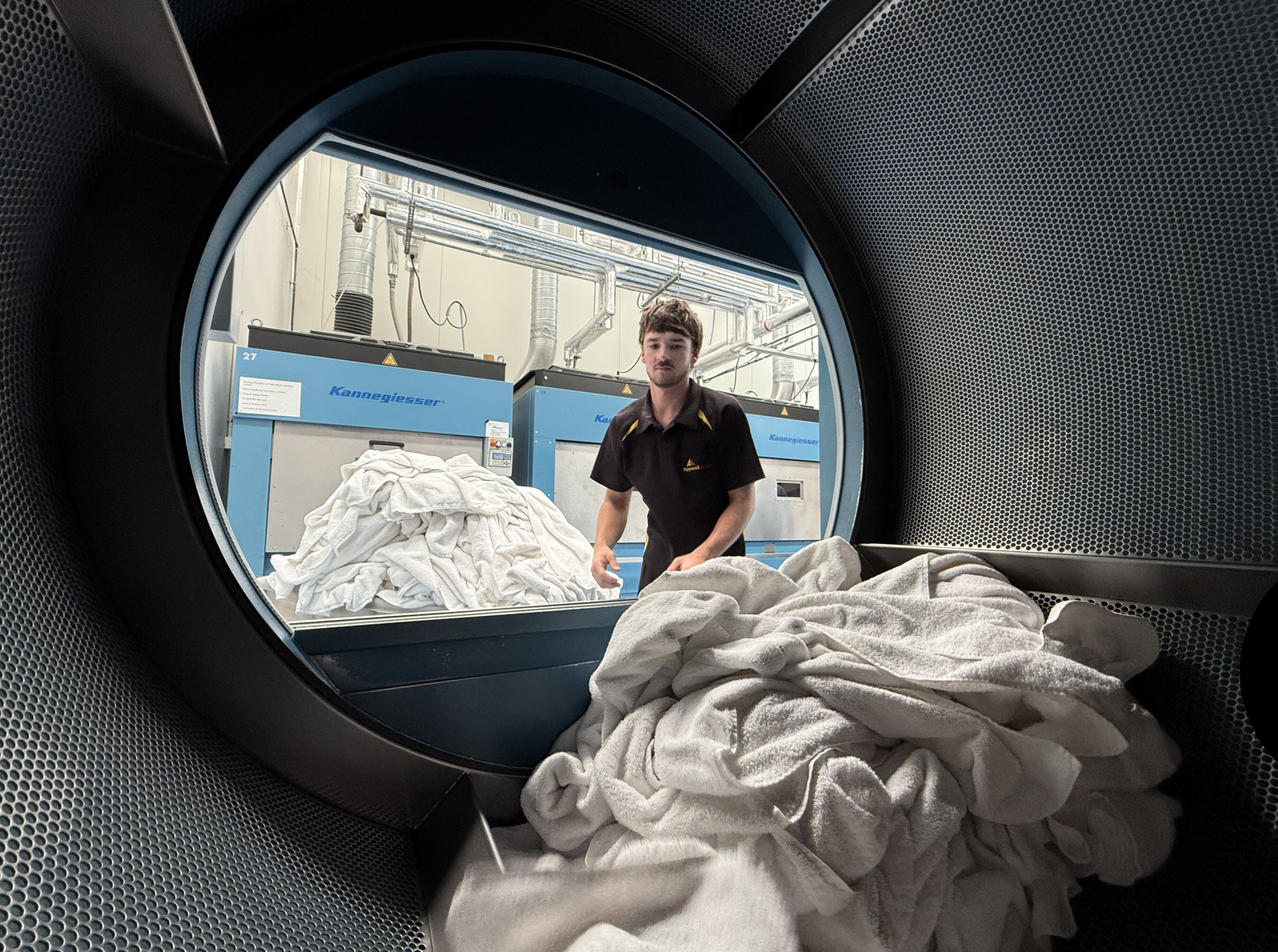 Looking through the door of a commercial dryer, a laundry worker is seen sorting white towels and linens outside the machine in a laundry facility.