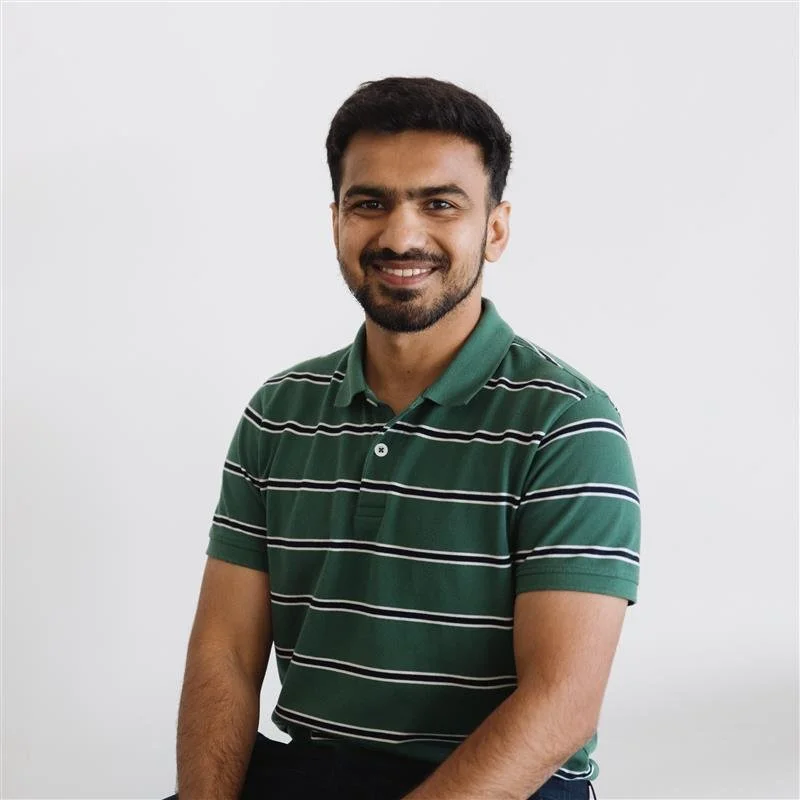 A man with dark hair and a beard smiling, wearing a green and black striped polo shirt, sitting against a plain white background.