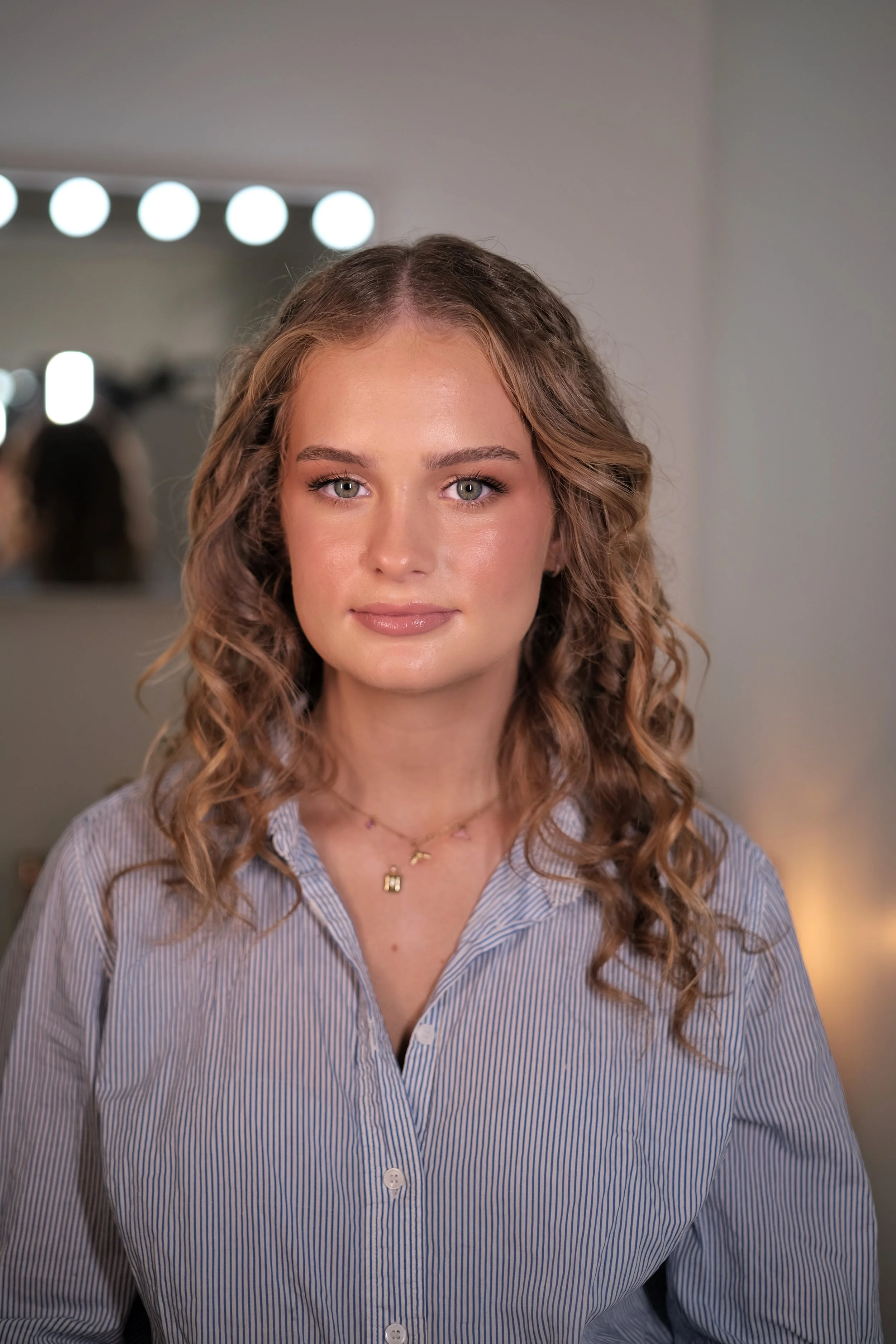 A young woman with wavy, shoulder-length hair, wearing a striped shirt, standing in a room with a large mirror and round lights in the background.