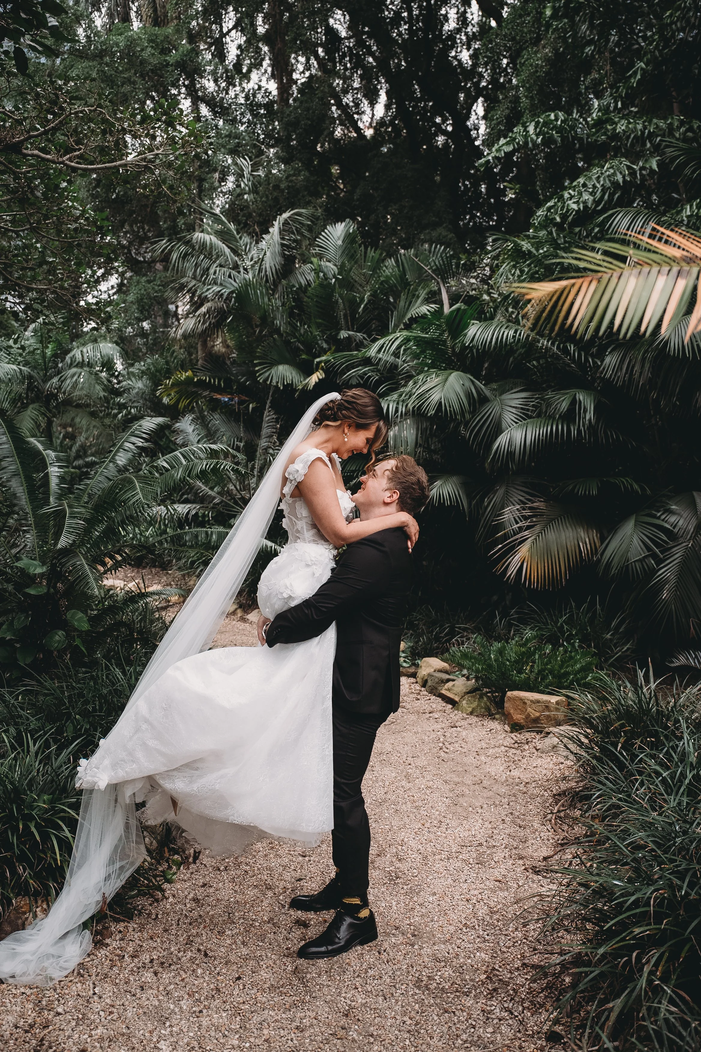A bride and groom in wedding attire in a lush, green garden, with the groom lifting the bride and both smiling at each other.