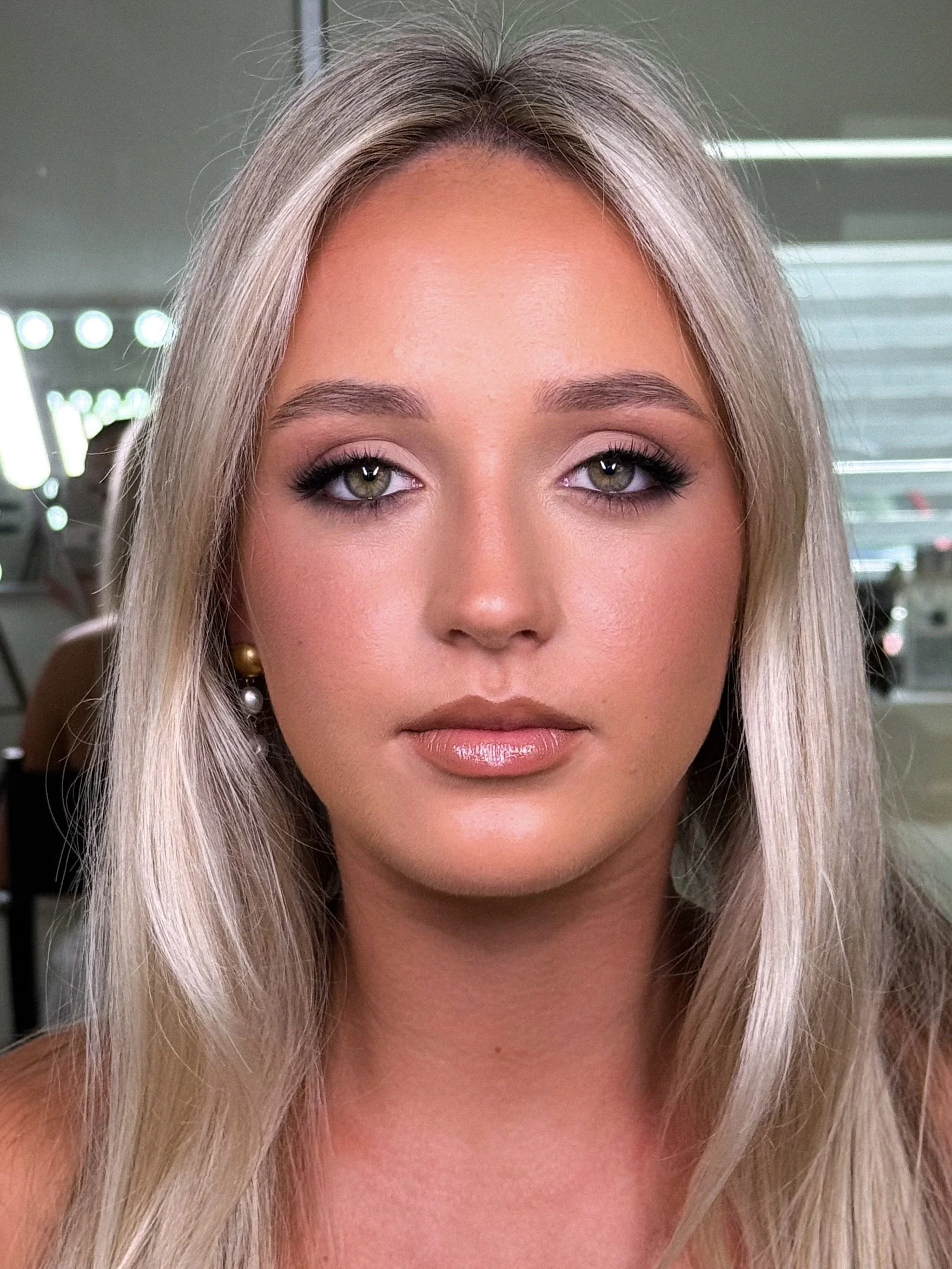 Close-up portrait of a young woman with long blonde hair, green eyes, and makeup, indoors with a blurred background.
