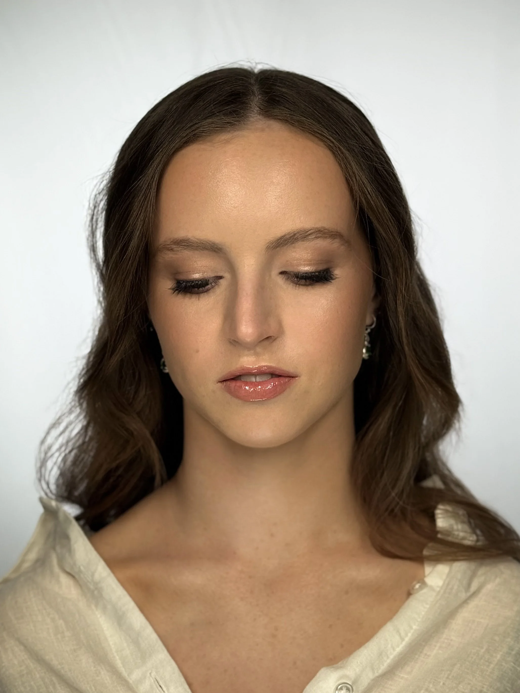 A young woman with brown hair styled in loose waves, wearing a light-colored top and earrings, with her eyes closed and a neutral expression.