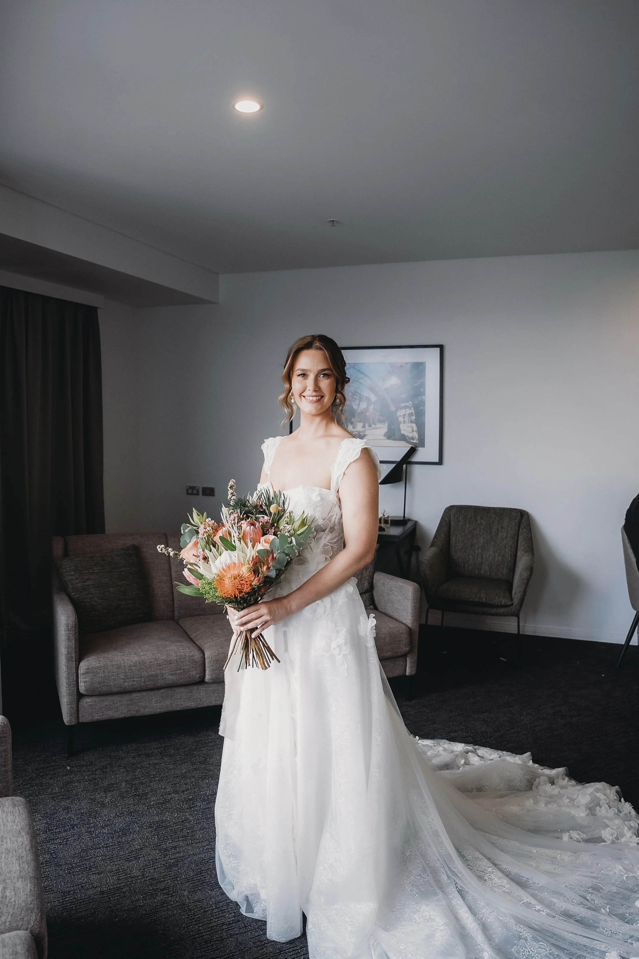 A smiling bride in a wedding dress holding a bouquet of flowers inside a room with modern furniture.