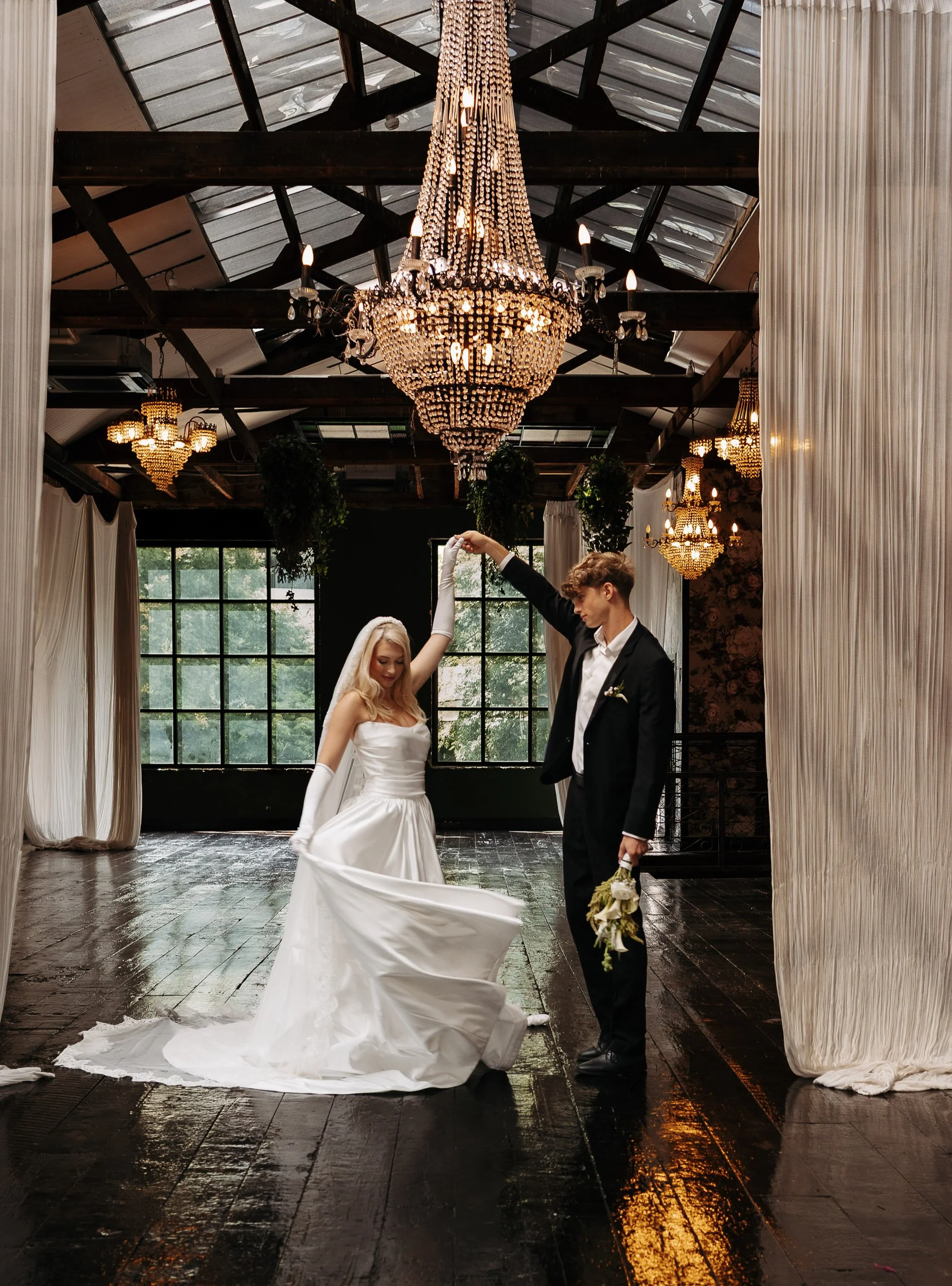 A bride and groom dancing in an elegant indoor venue with large chandeliers and dark wooden floors, with the bride wearing a strapless white gown and the groom in a black suit holding a bouquet.