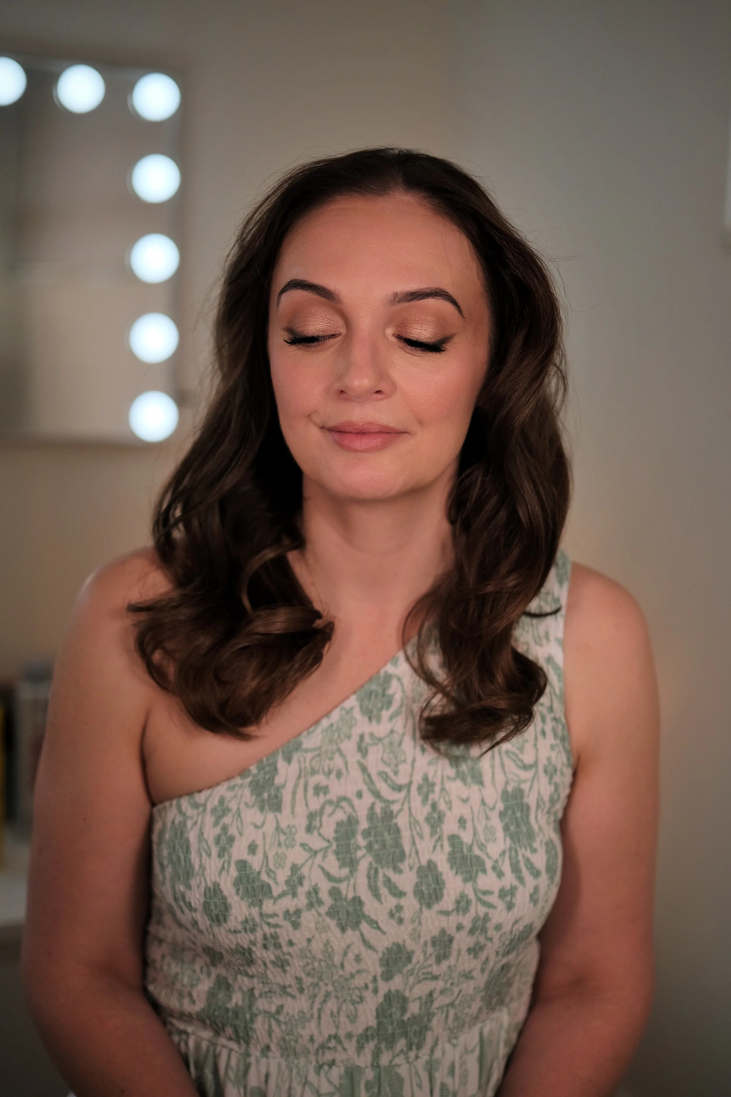 A woman with long brown hair and closed eyes, smiling gently, wearing a sleeveless floral dress, in front of a mirror with round lights.