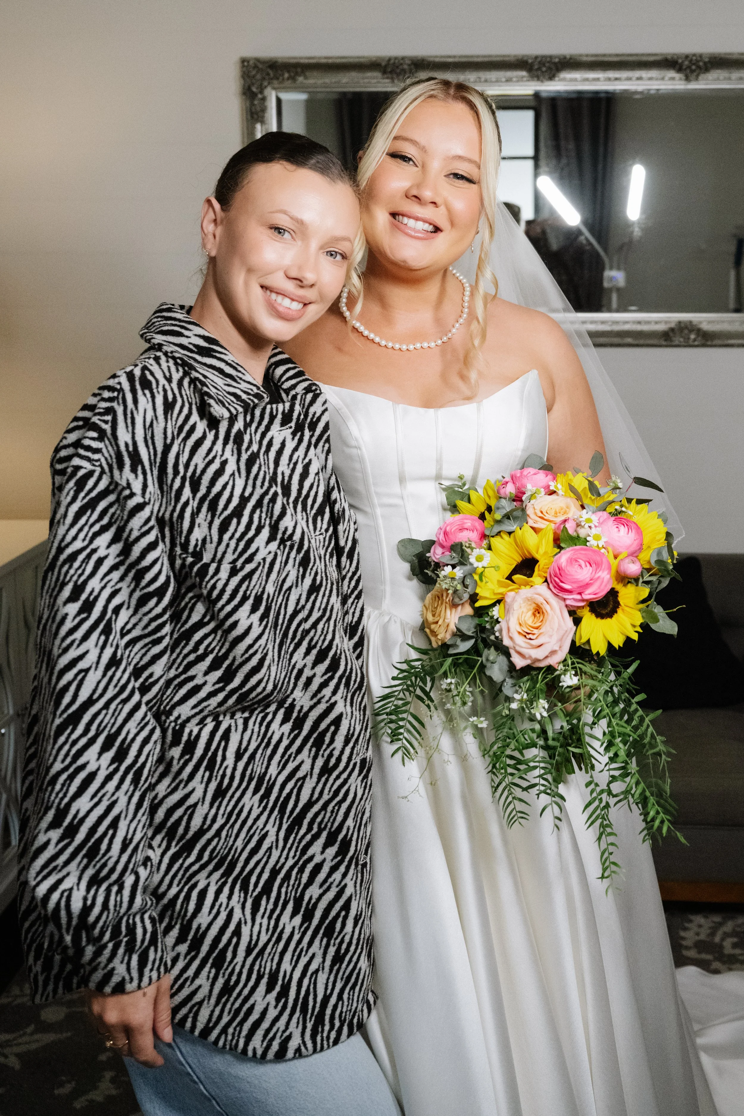 Two women, one in a wedding dress holding a bouquet, and the other in a zebra print coat, smiling together in a cozy room.