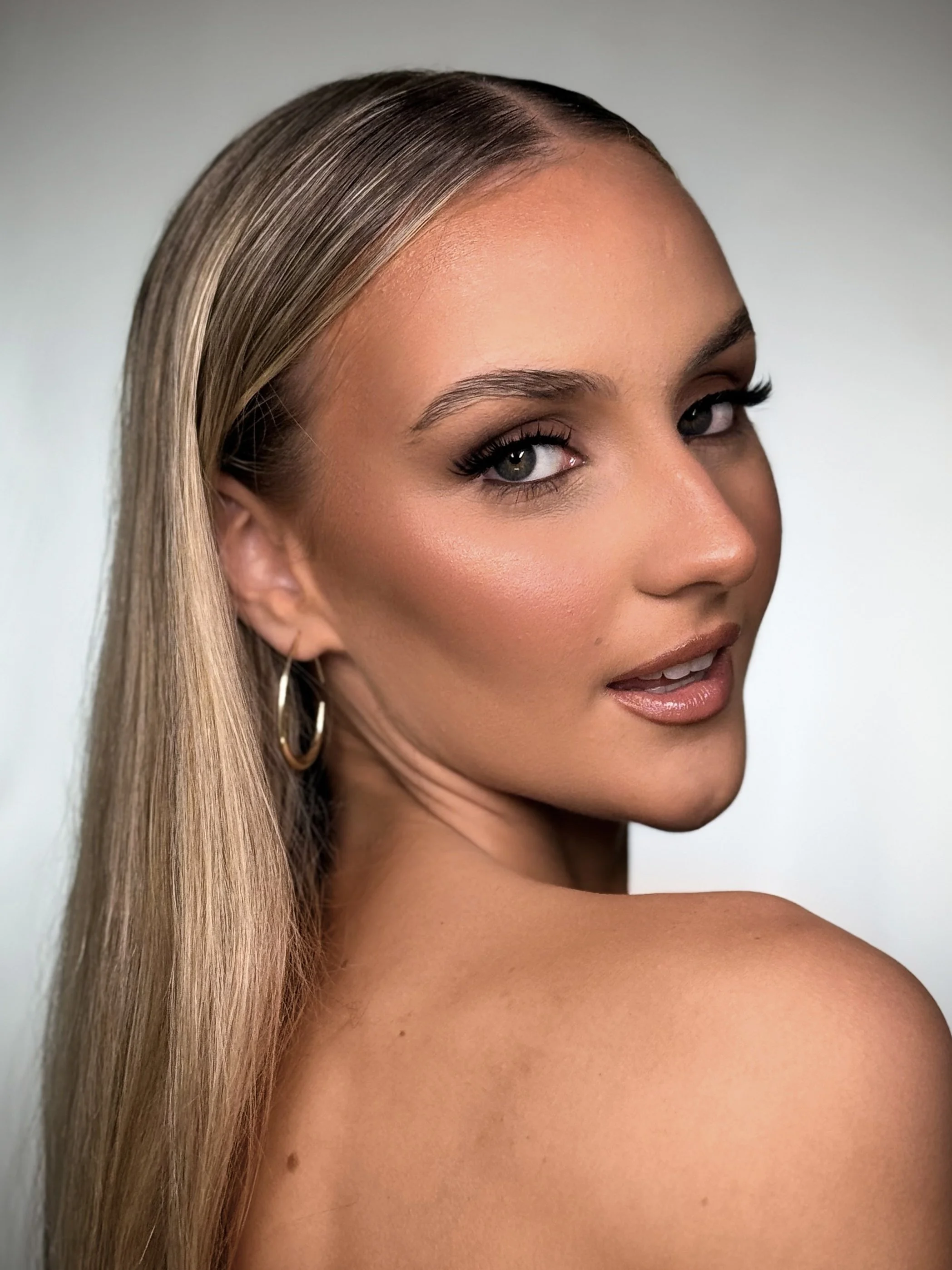 Close-up portrait of a woman with blonde hair, wearing hoop earrings, with makeup and a confident expression, looking over her shoulder.