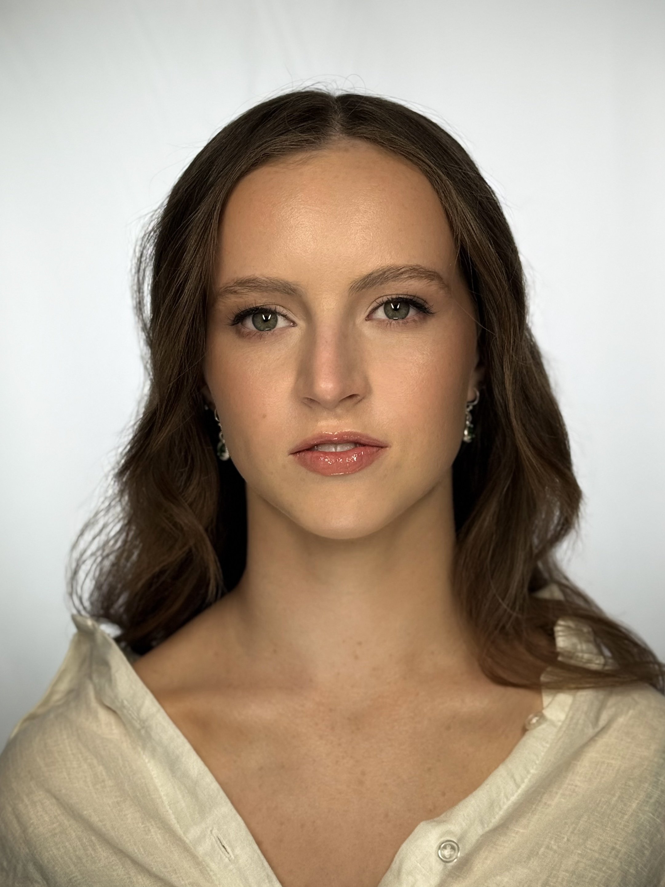 Close-up portrait of a young woman with brown wavy hair, green eyes, and wearing earrings and a light-colored top, against a plain background.