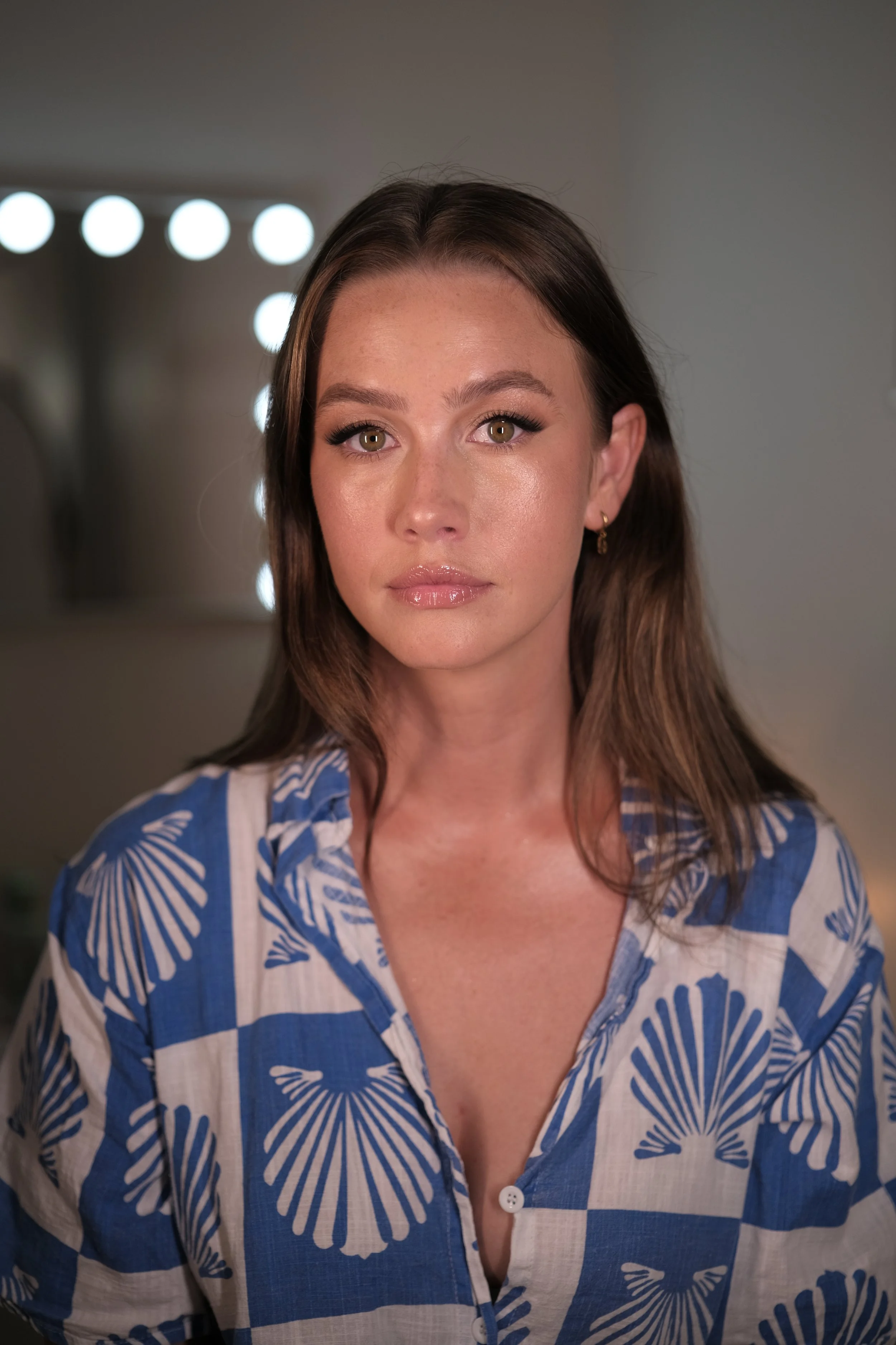 A young woman with long brown hair wearing a blue and white patterned shirt, looking directly at the camera.