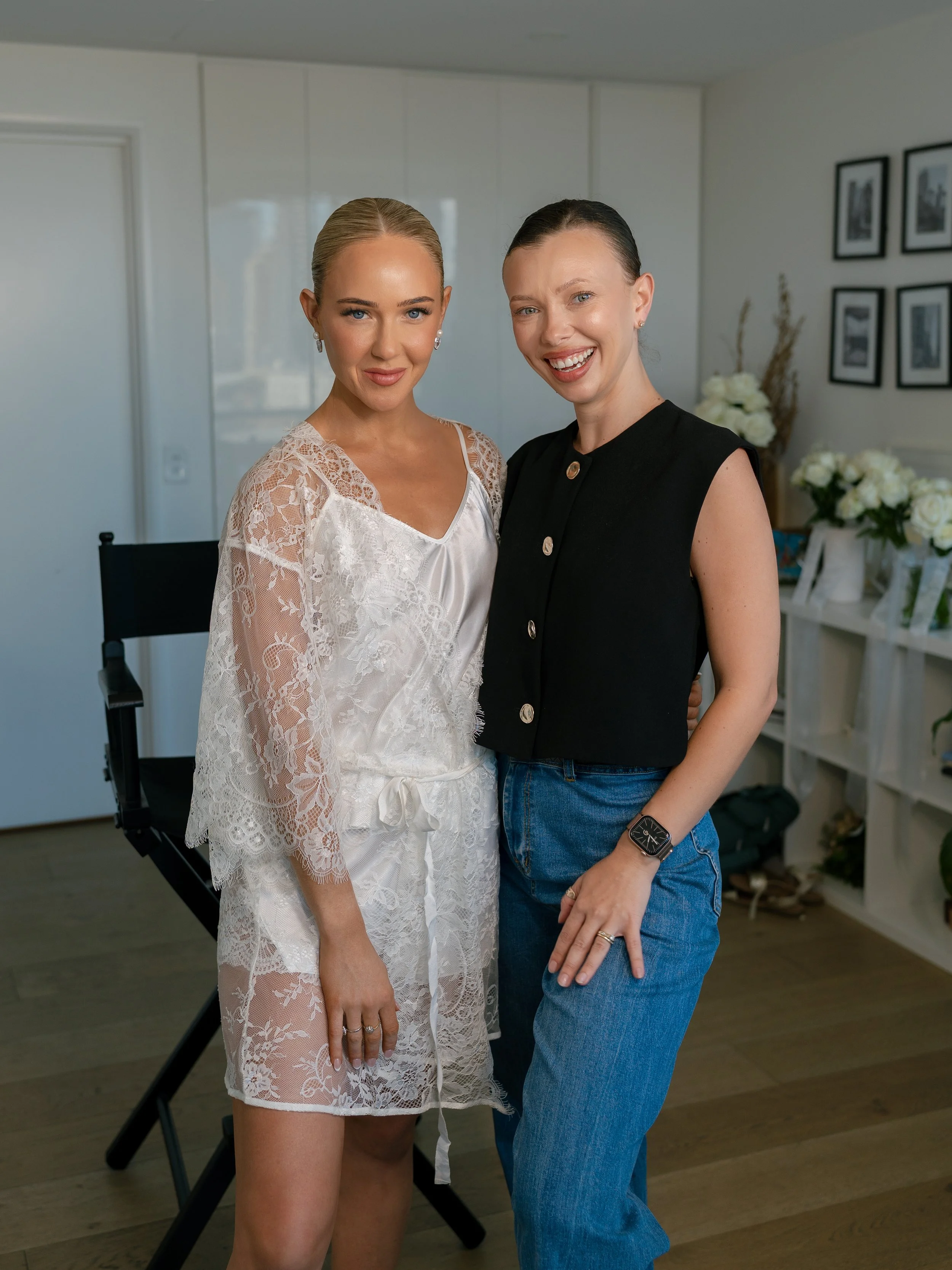 Two women smiling and posing together indoors; one is in a white lace robe, and the other is in a black sleeveless top and blue jeans. The background features framed photos and white floral arrangements.