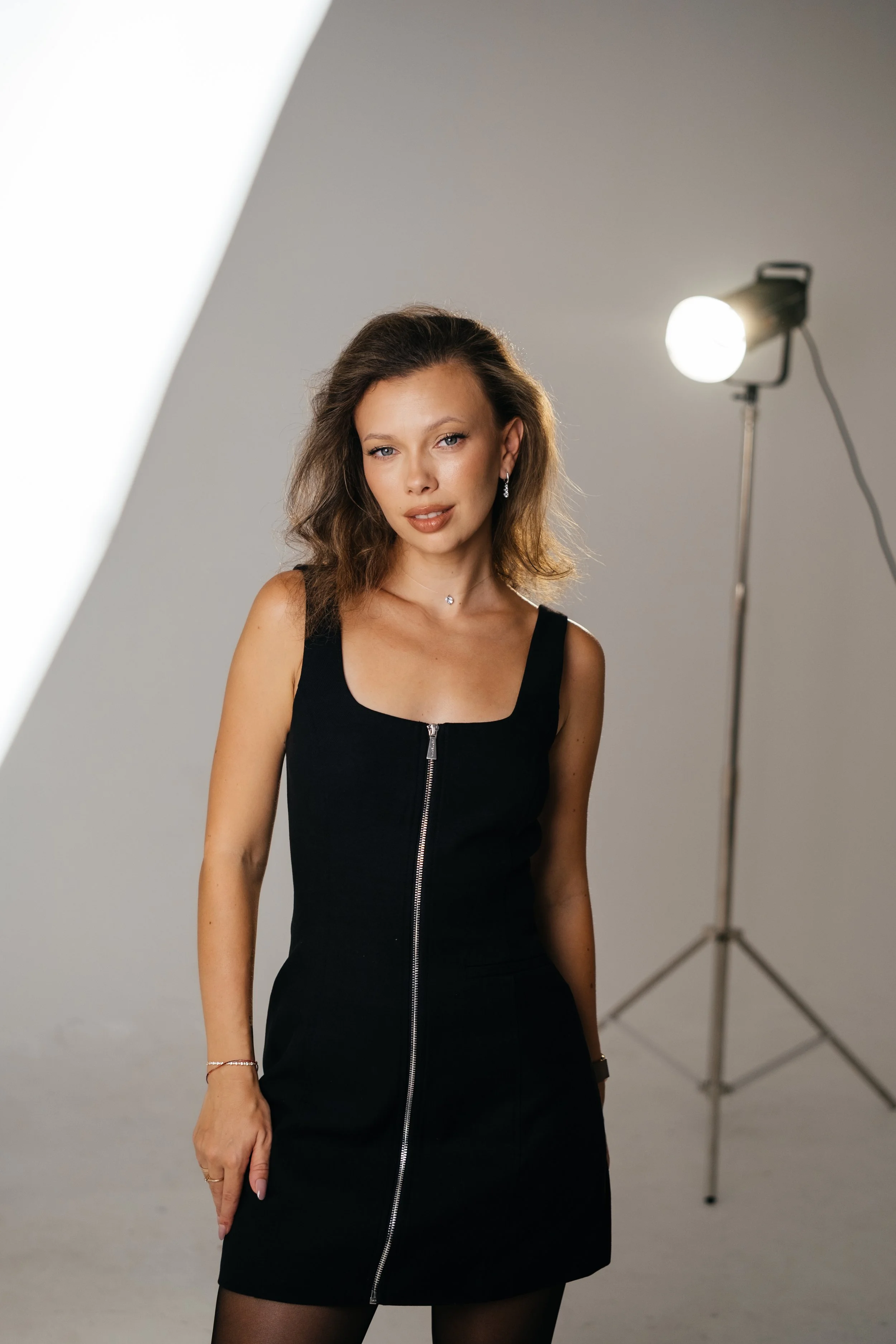 Portrait of a young woman with wavy brown hair wearing a black sleeveless dress with a front zipper, standing in a photography studio with professional lighting equipment.