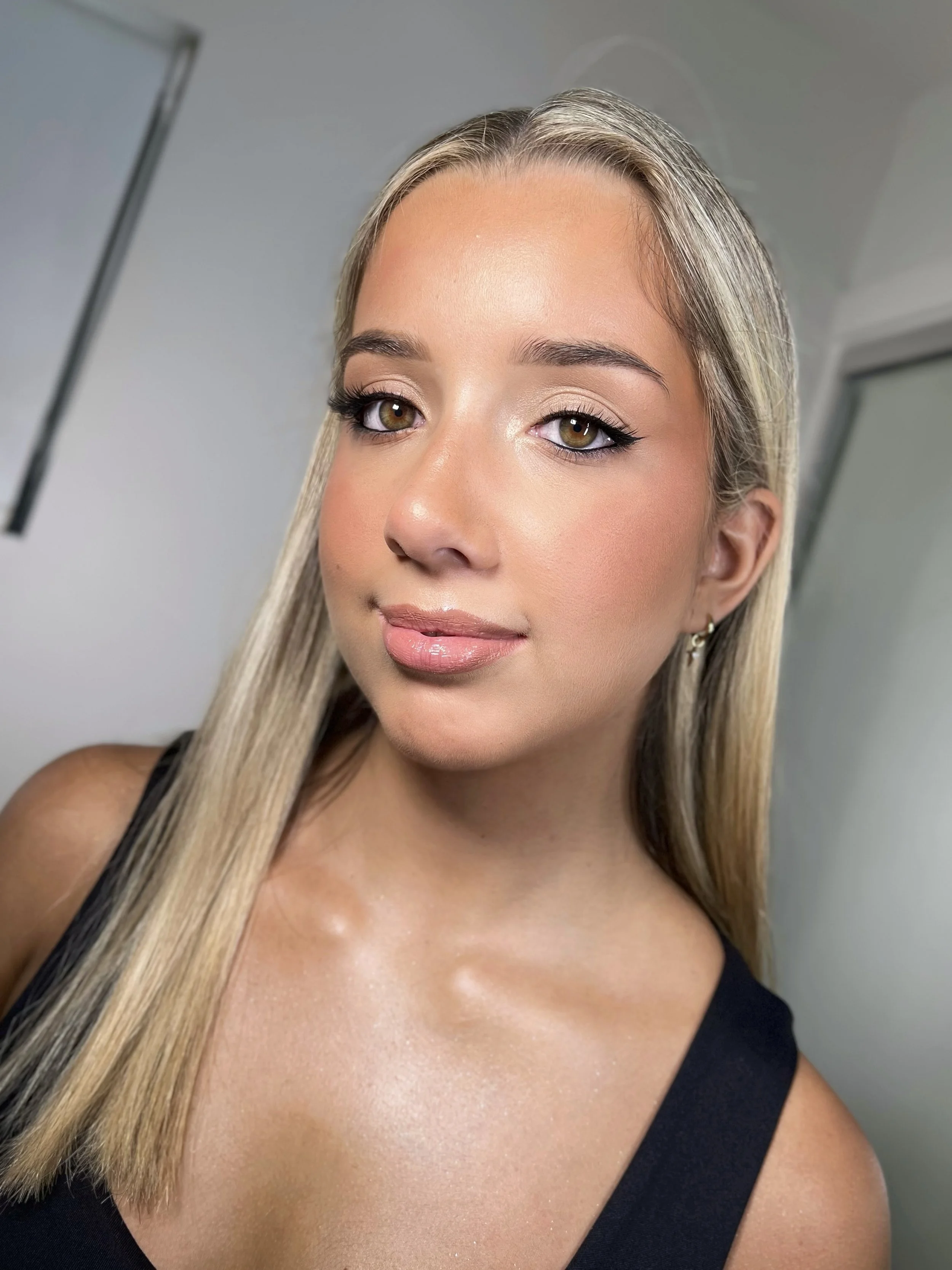 Close-up of a young woman with long blonde hair, wearing makeup, earrings, and a black sleeveless top, smiling softly.
