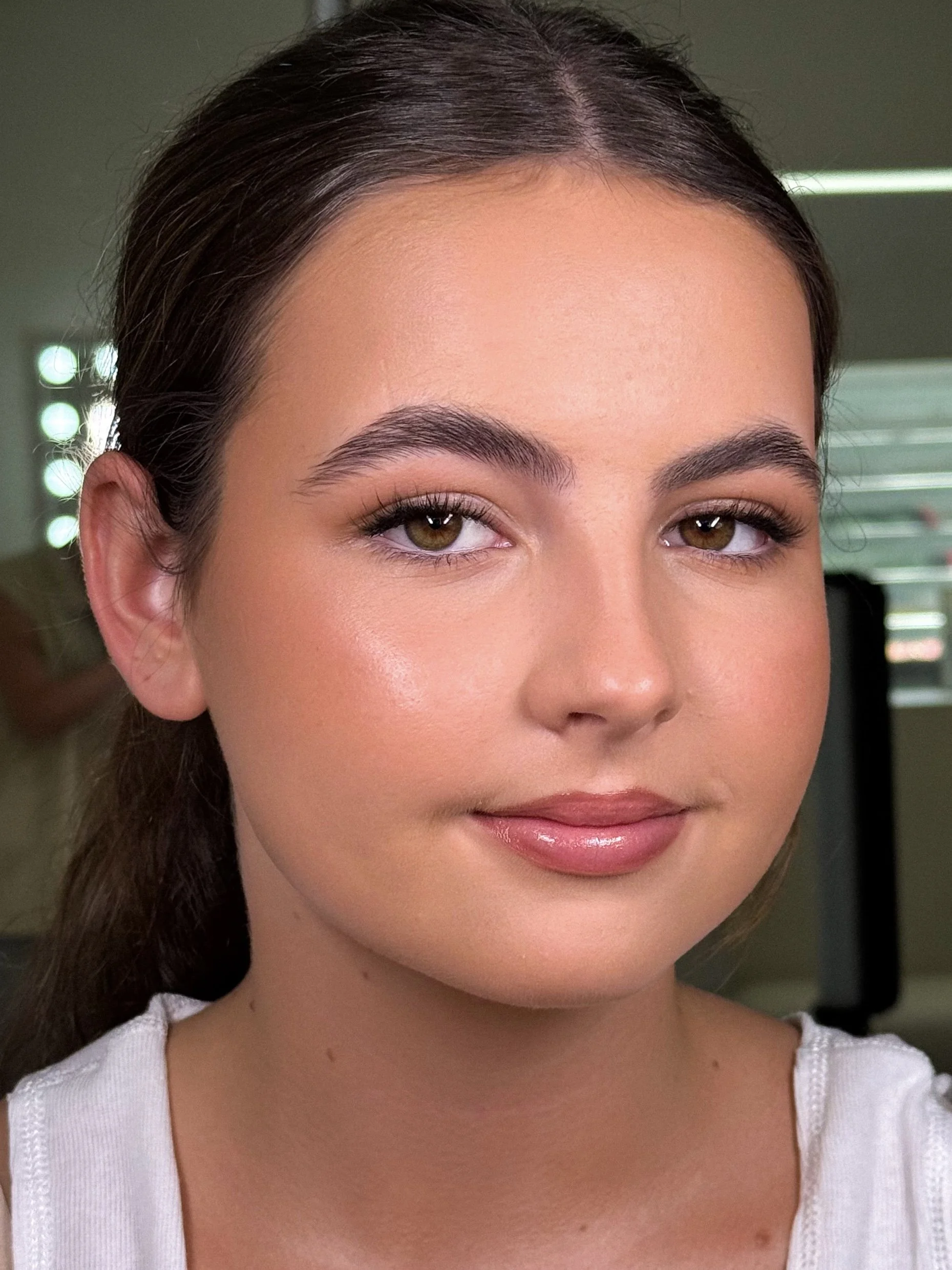 Close-up of a young woman with brown hair, light makeup, and a white shirt, smiling softly in indoor natural lighting.